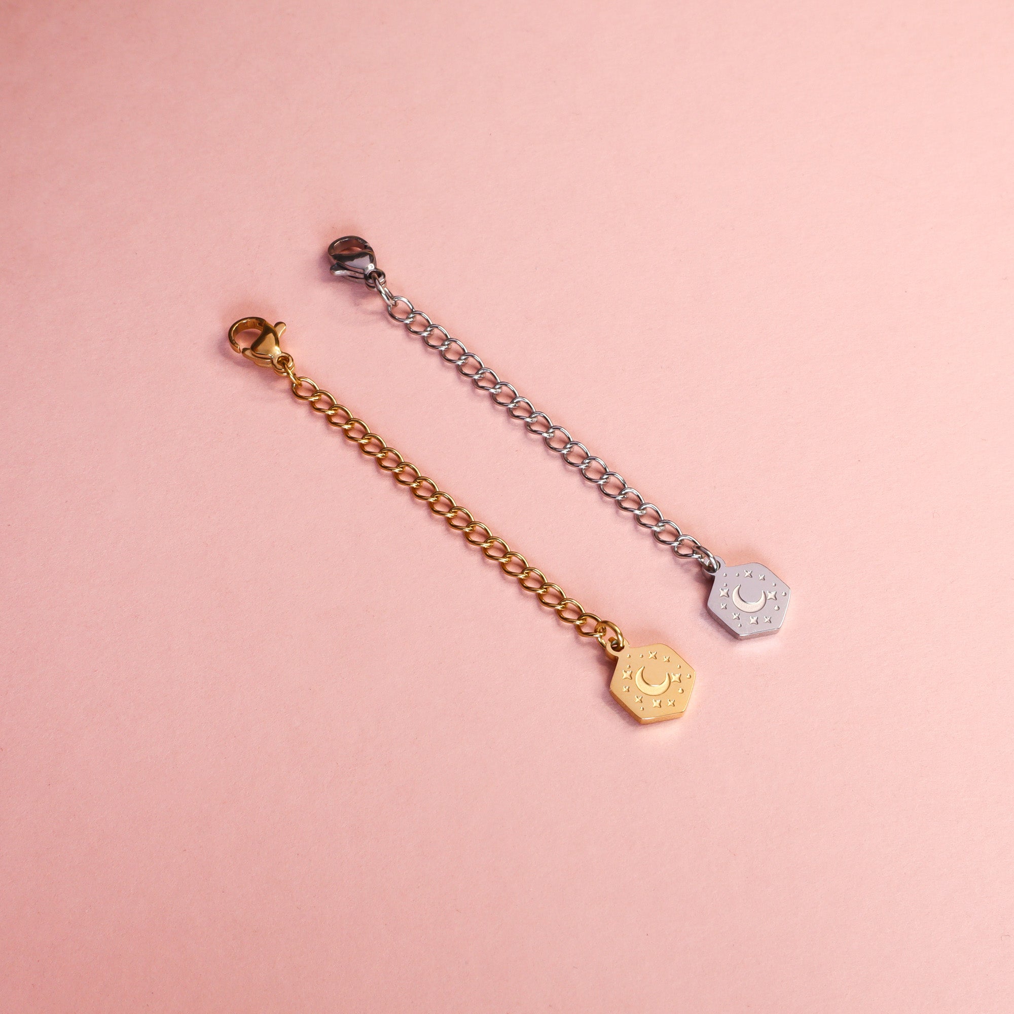 Two necklace extenders, one gold and one silver taken on a light pink background. The extenders feature a small hexagon with etched moon and star details.
