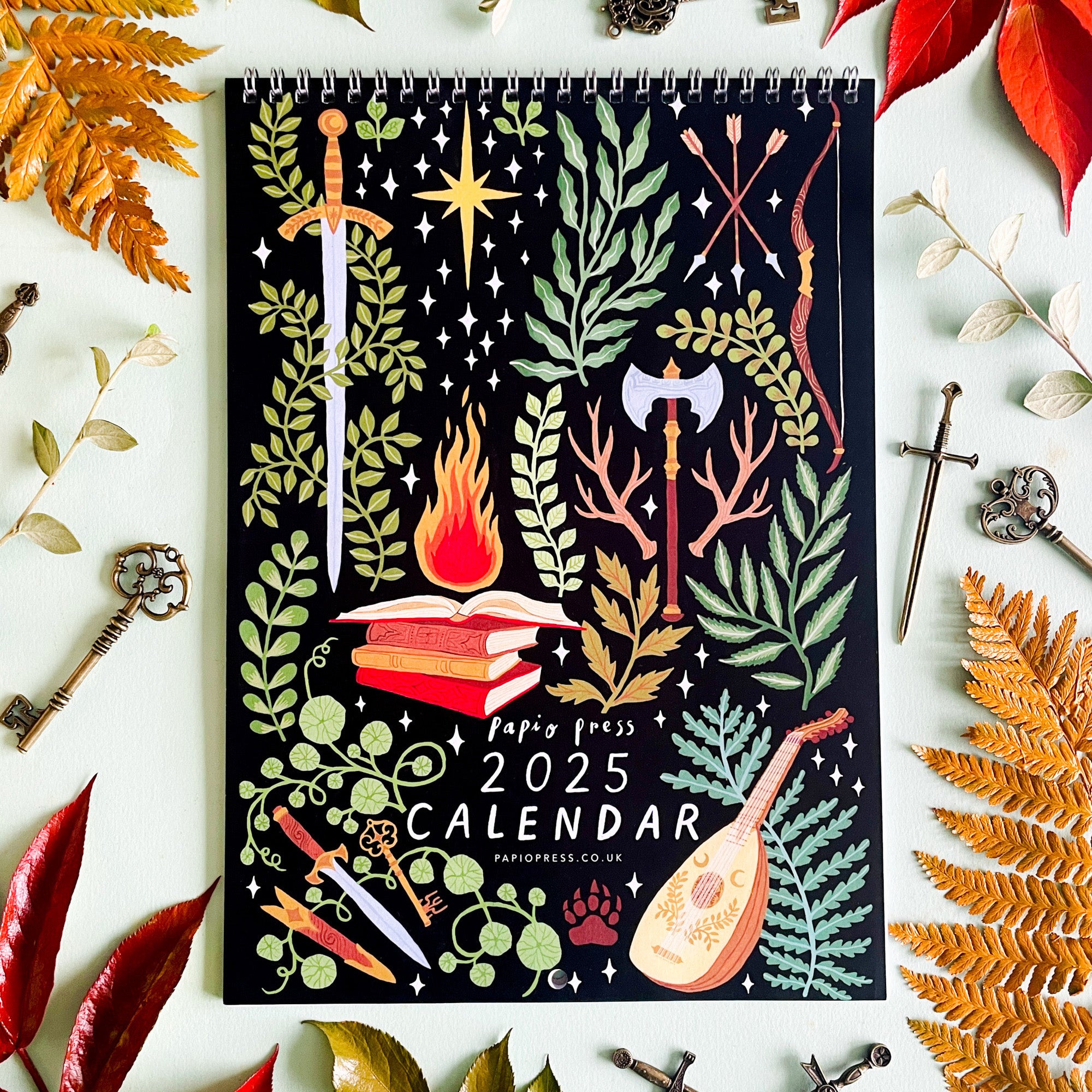 The 2025 Papio Press Fantasy Calendar features an illustrated cover with swords, keys, a lute, axe, magical books aflame, and autumn leaves in the enchanting style of Harry & Zanna Goldhawk for Papio Press.