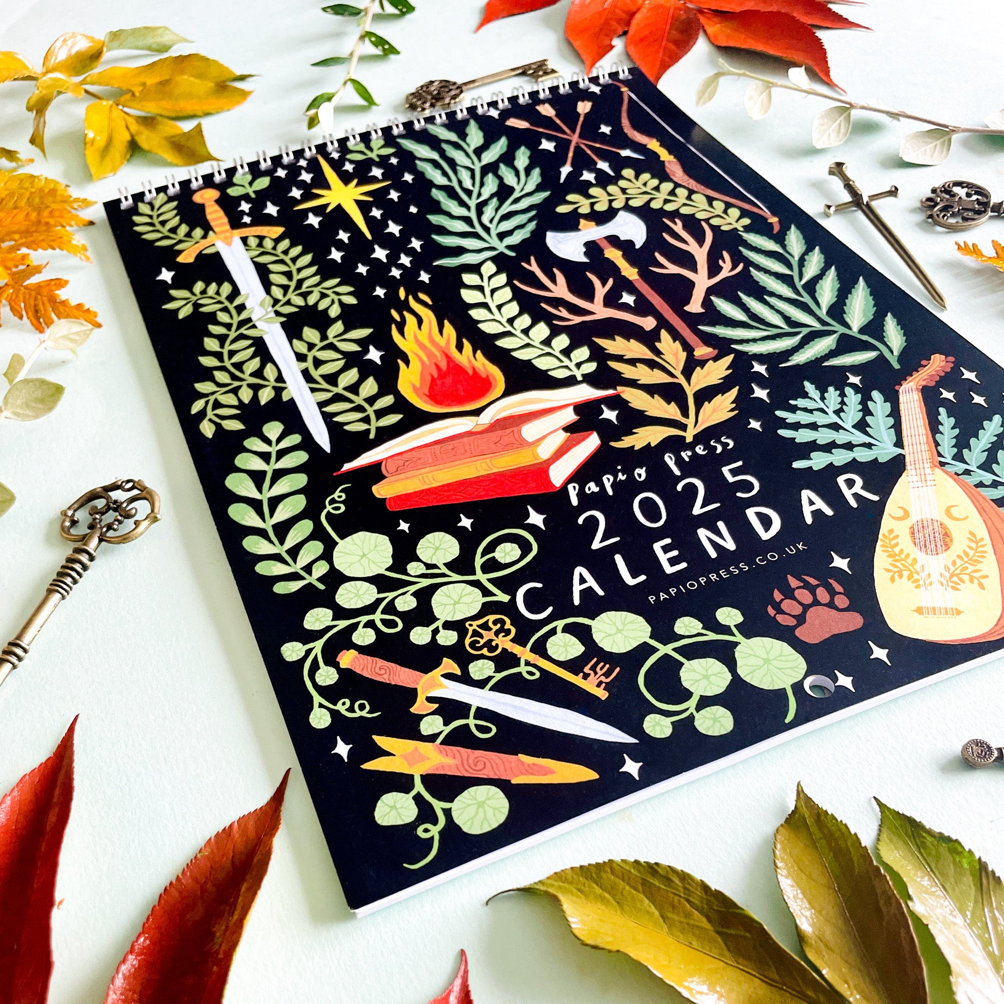 The 2025 Papio Press Fantasy Calendar features magical Harry & Zanna Goldhawk illustrations of plants, swords, books, and more. Surrounded by autumn leaves, keys, and a feather, this Papio Press calendar adds charm to any space.