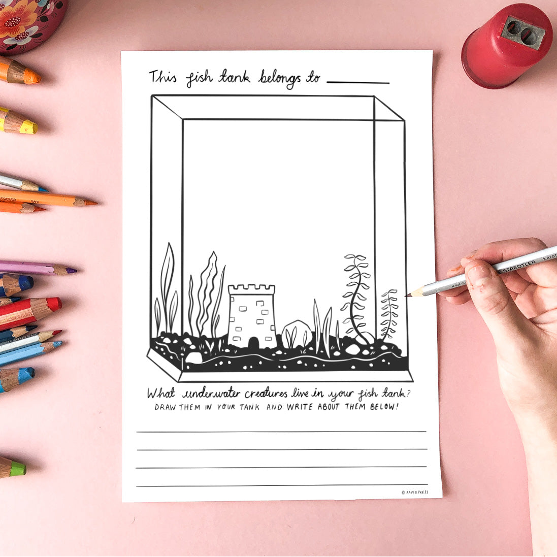 A hand holds a pencil above a fish tank-themed worksheet with space to draw underwater creatures and write about them. Colored pencils and a pencil sharpener are scattered on a pink surface.