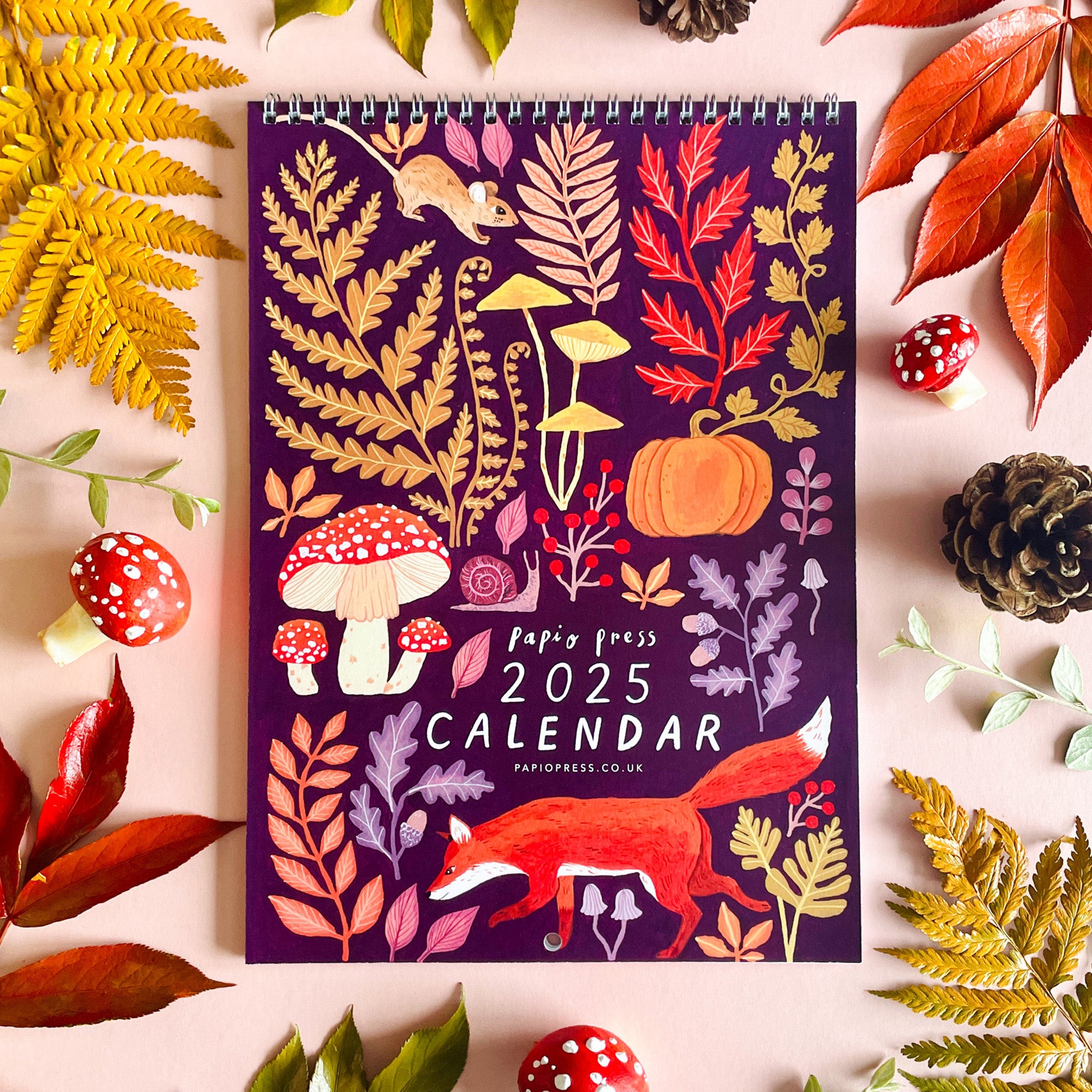The 2025 Papio Press Florals & Animals Calendar features autumn woodland illustrations—fox, mushrooms, pumpkin, leaves, and ferns—set on a peach background with real leaves and pinecones for a charming seasonal look.