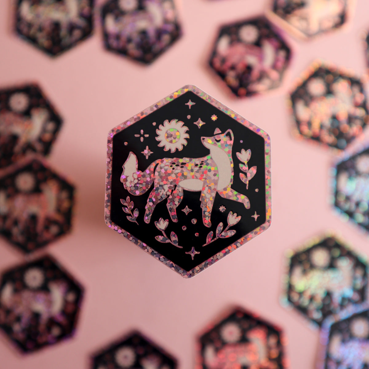 A hexagonal sticker or patch featuring a glittery pink fox on a black background with a holographic glitter border, displayed against a pink backdrop with other blurred stickers.