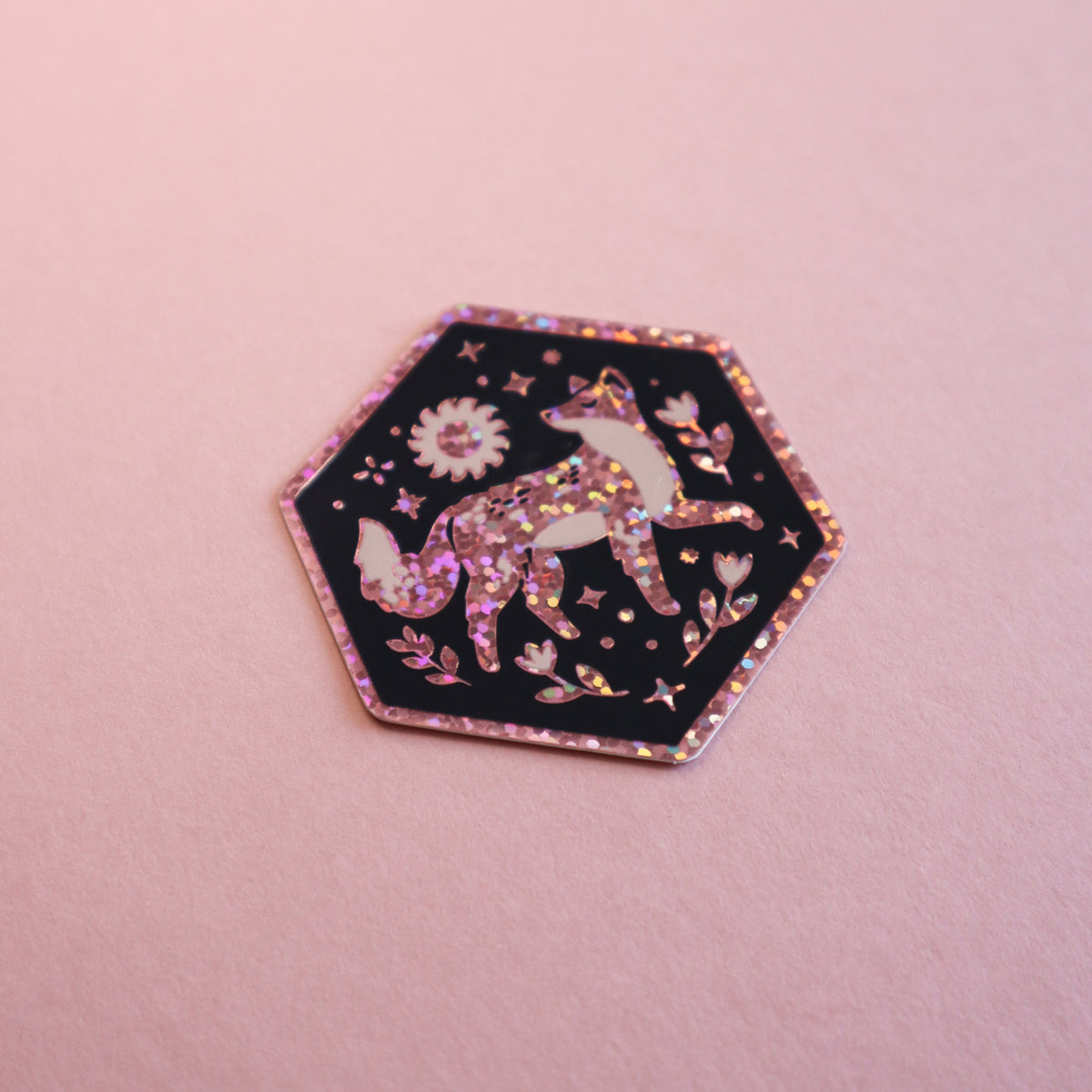 A hexagonal enamel pin featuring a glittery pink fox on a black background with floral and star details, set against a solid pink background.