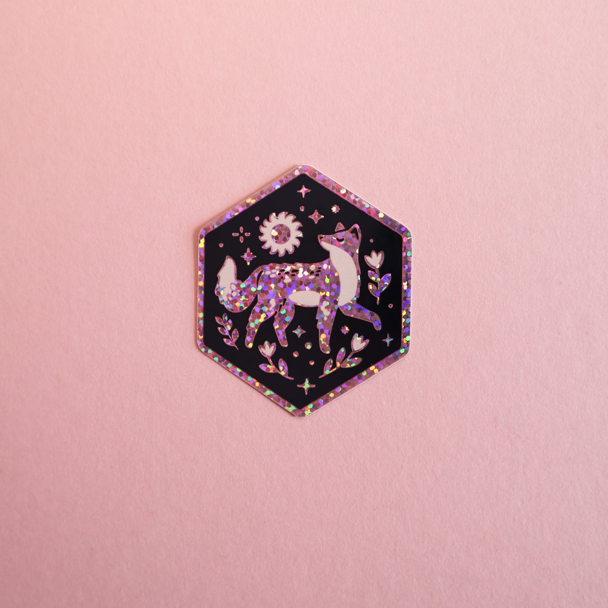A hexagonal enamel pin featuring a glittery purple fox on a black background with pink and holographic accents, displayed on a solid pink surface.