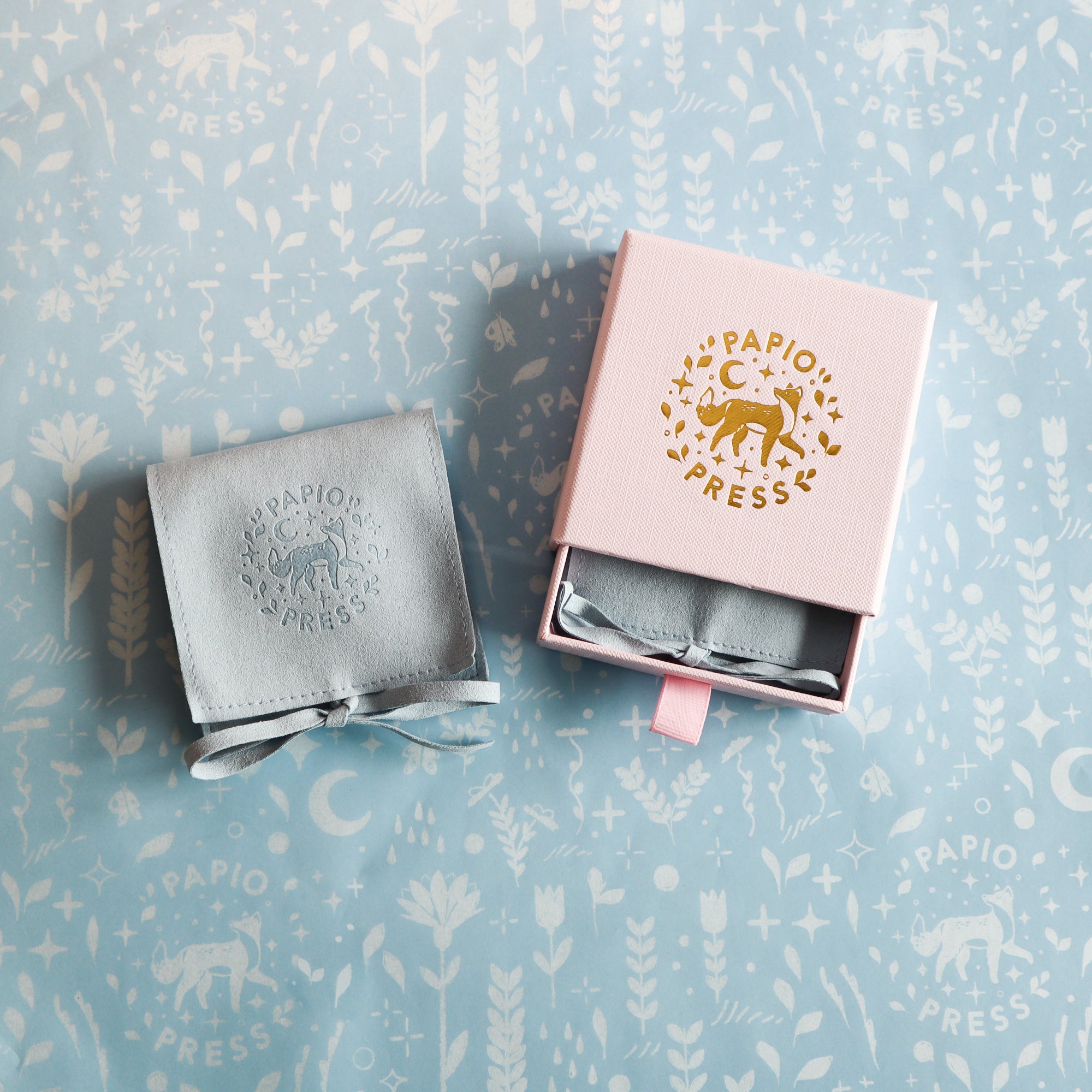 A light blue Papio Press pouch with a gold logo sits beside a pink gift box—both ideal for presenting the Silver Fox Pendant—on matching blue patterned paper featuring animal and botanical designs.
