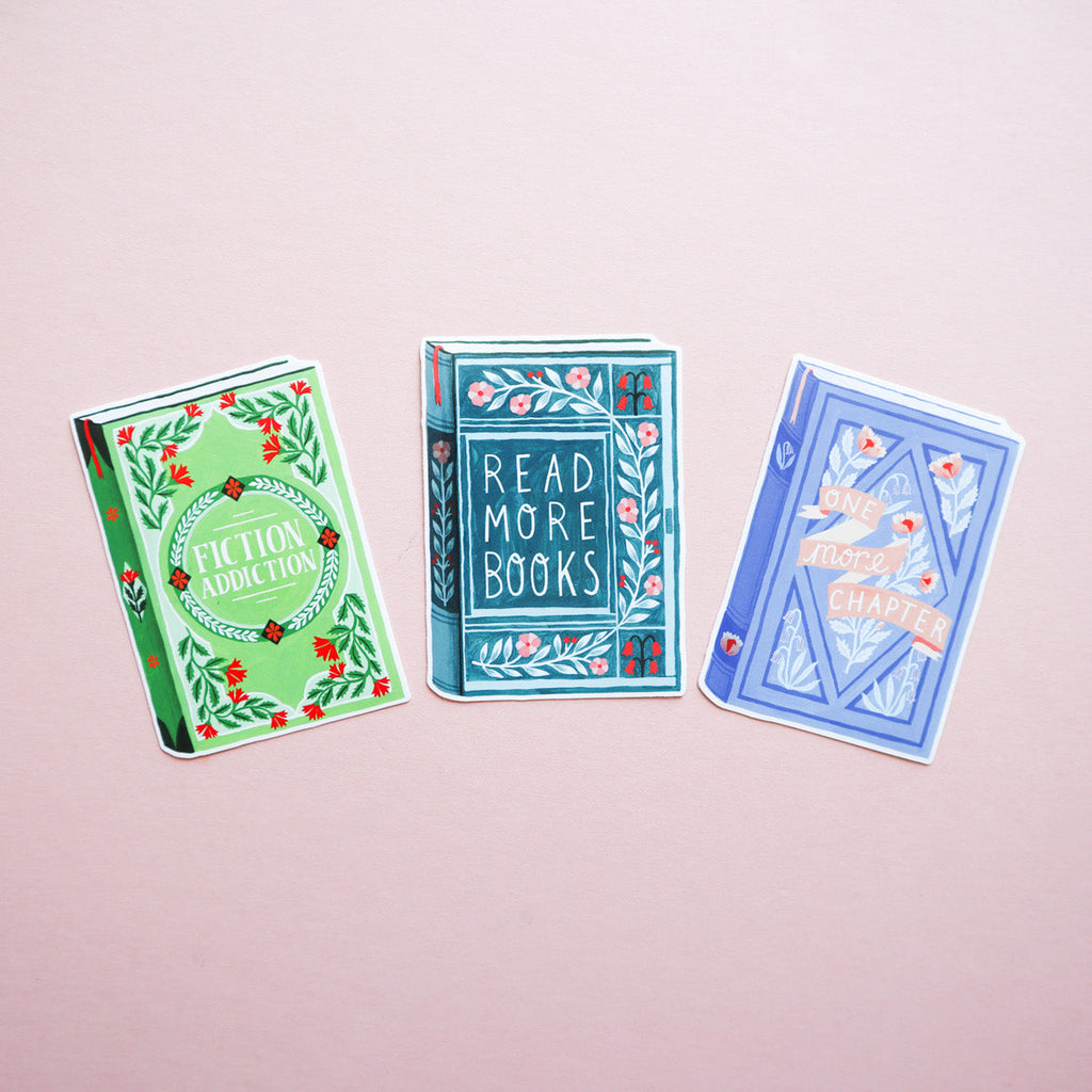 A picture of three book stickers, one says 'Fiction Addiction', 'Read More Books' and 'One more Chapter'. The photo is taken on a pink background.
