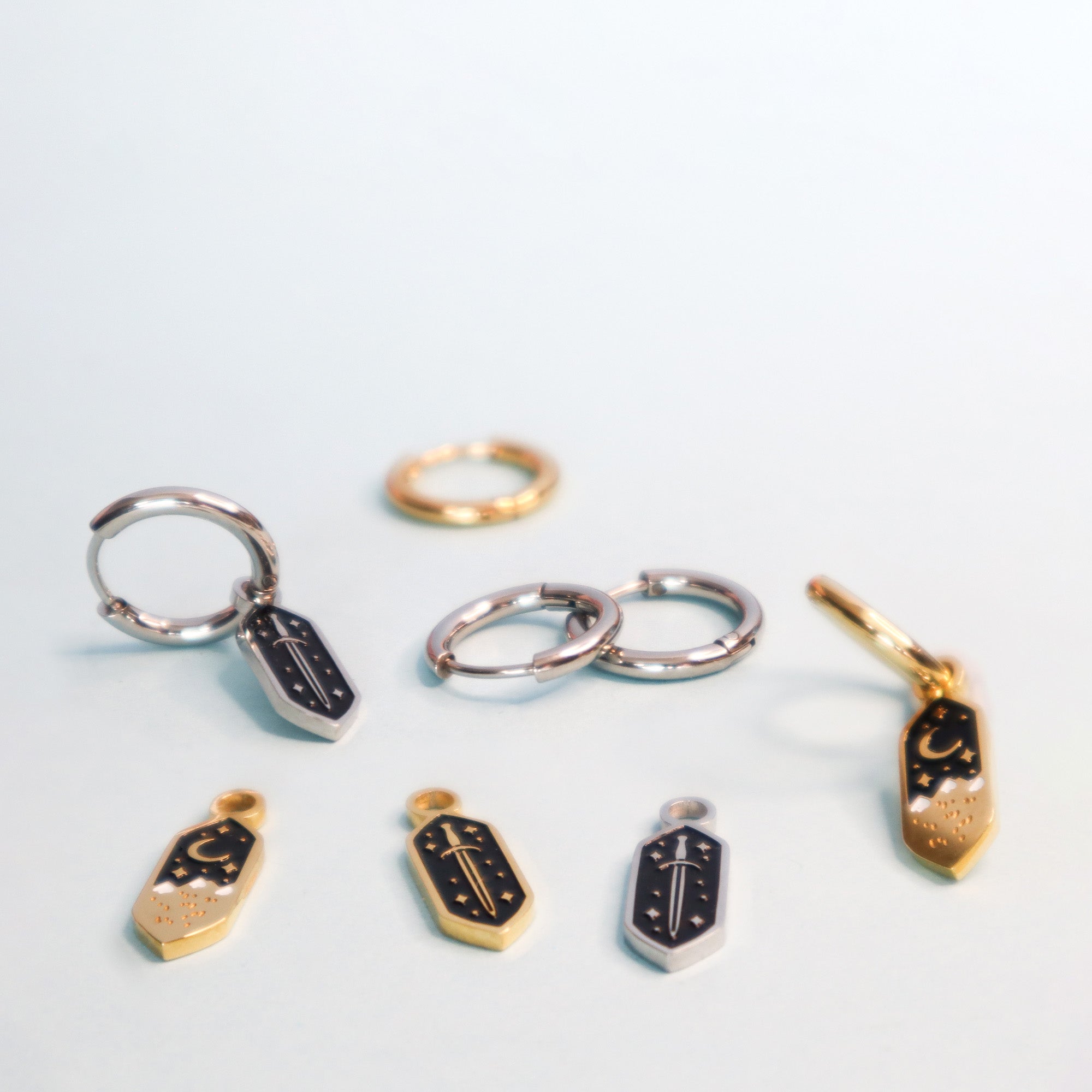 Papio Press Mini Mountains Hoop Earrings feature gold-plated hoops with black and gold geometric charms, including enamel star and sword designs. Loose matching charms complete this striking collection, arranged on a light surface.