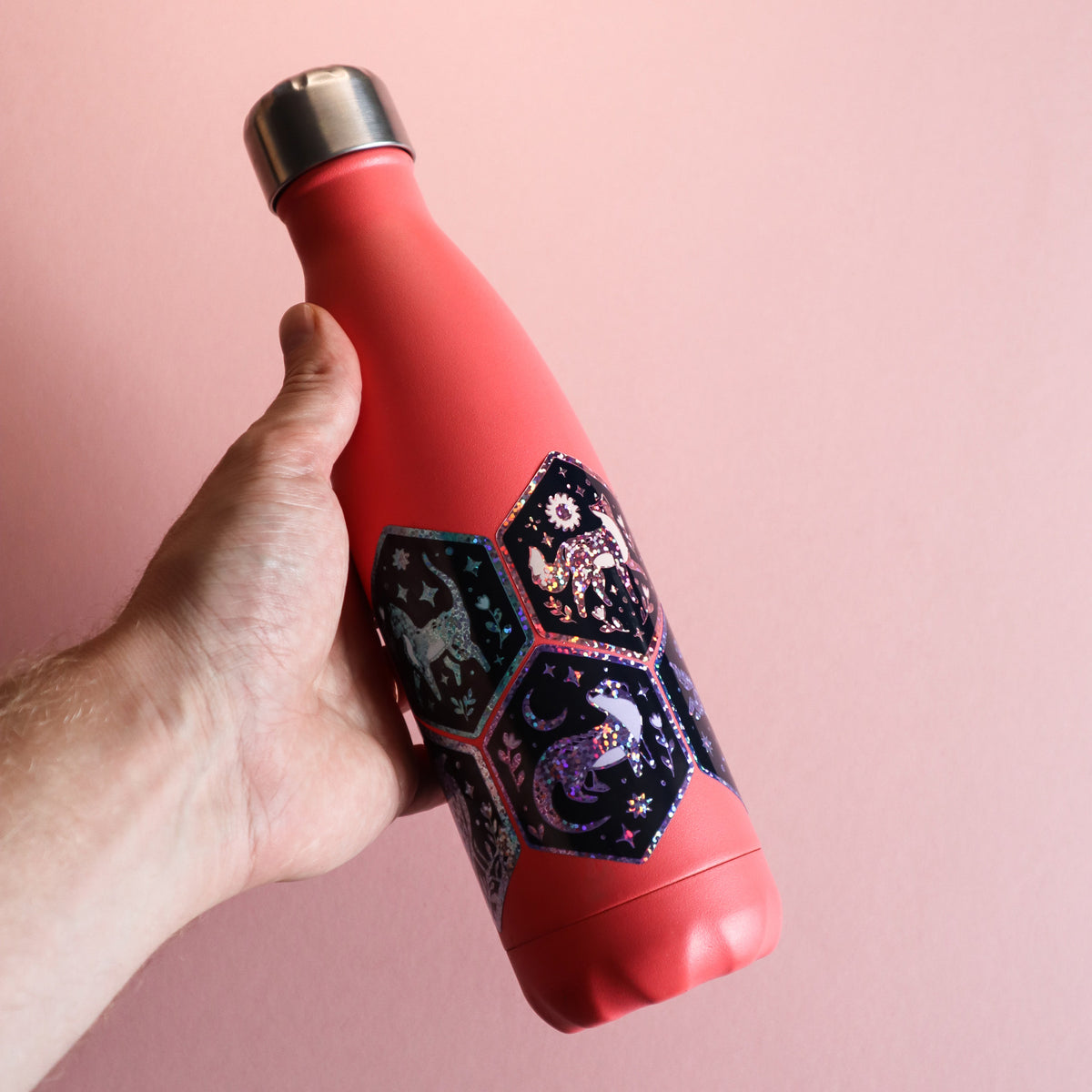 A hand holding a coral red insulated water bottle with a metallic silver cap and a decorative black hexagonal pattern featuring glittery cat and otter illustrations, set against a solid pink background.