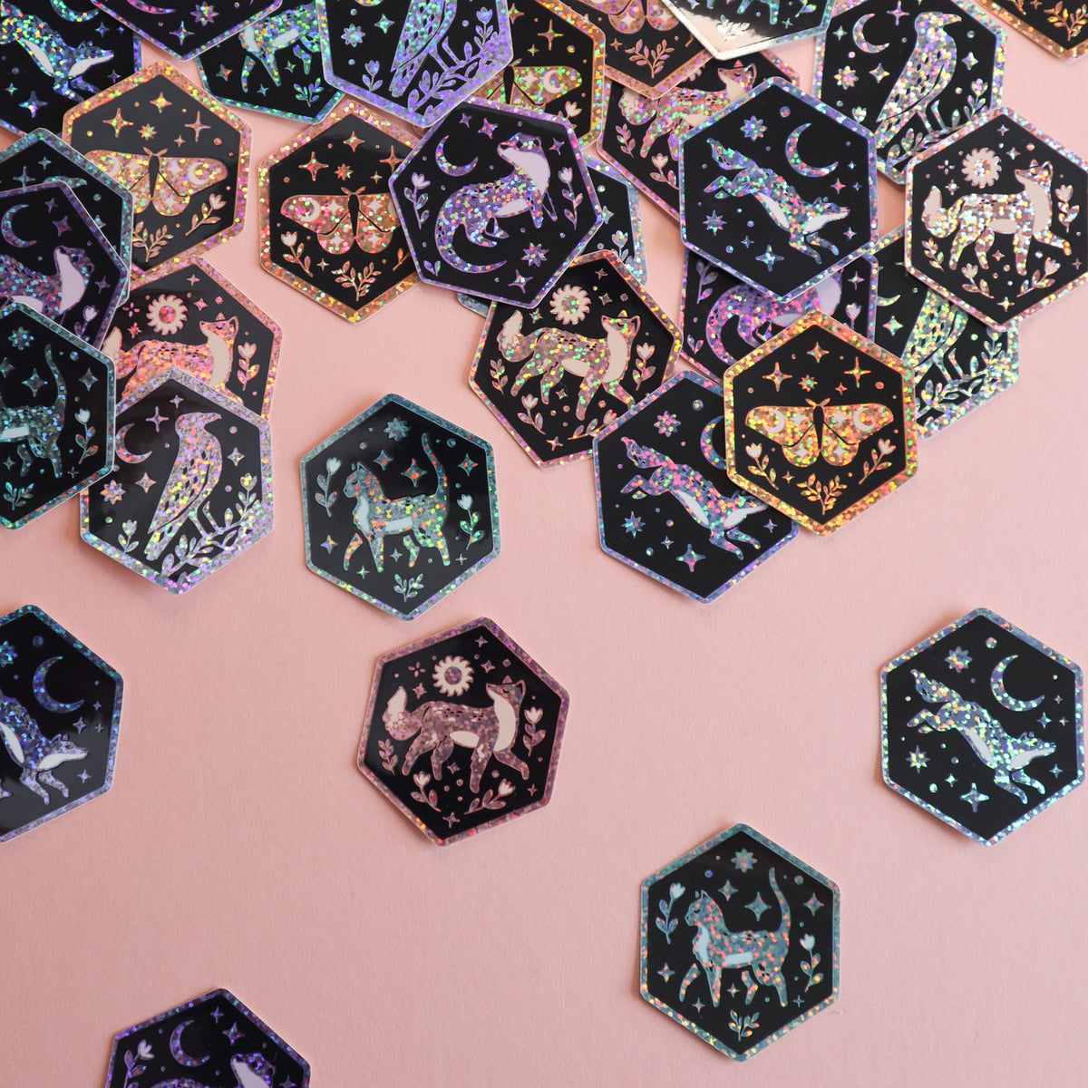 Hexagonal stickers featuring celestial and animal motifs in an iridescent holographic finish, scattered on a solid pink background.