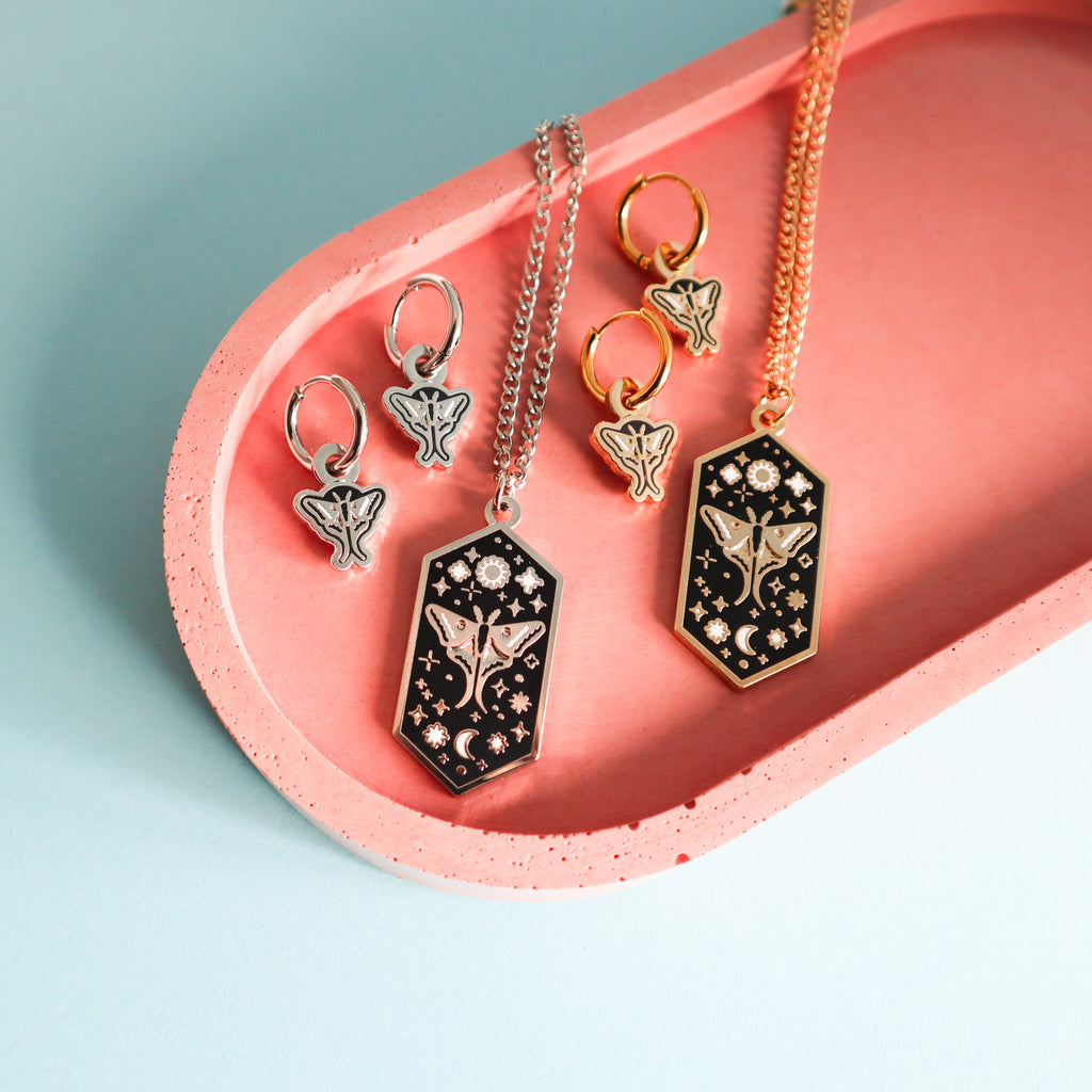 A selection of gold and silver moth earrings and necklaces on a pink concrete tray.