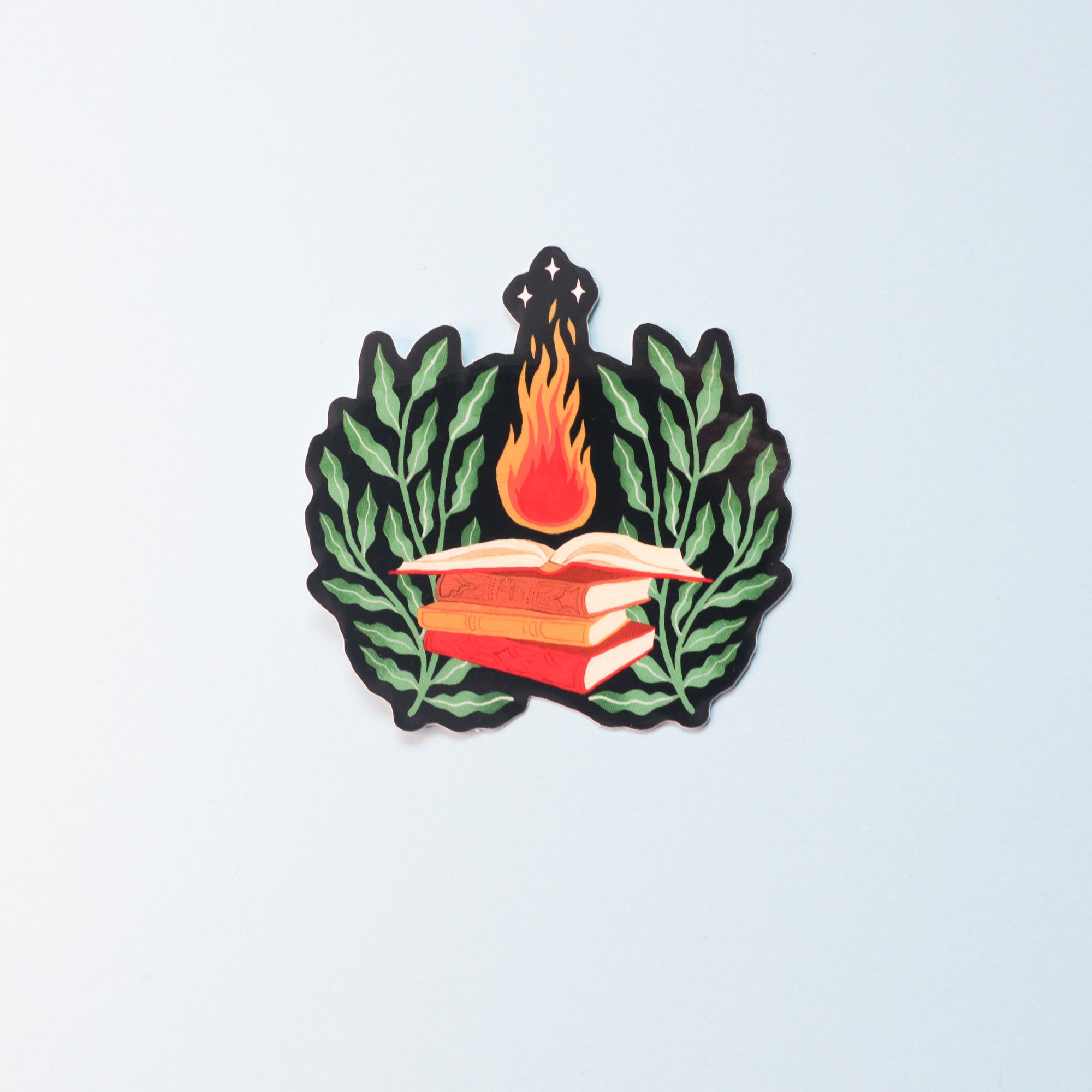 The Sorcerer Sticker by Papio Press features two red books stacked before green leafy branches, with an orange flame and three stars above them. This illustrated vinyl sticker has a glossy finish and UV protection for lasting vibrance.