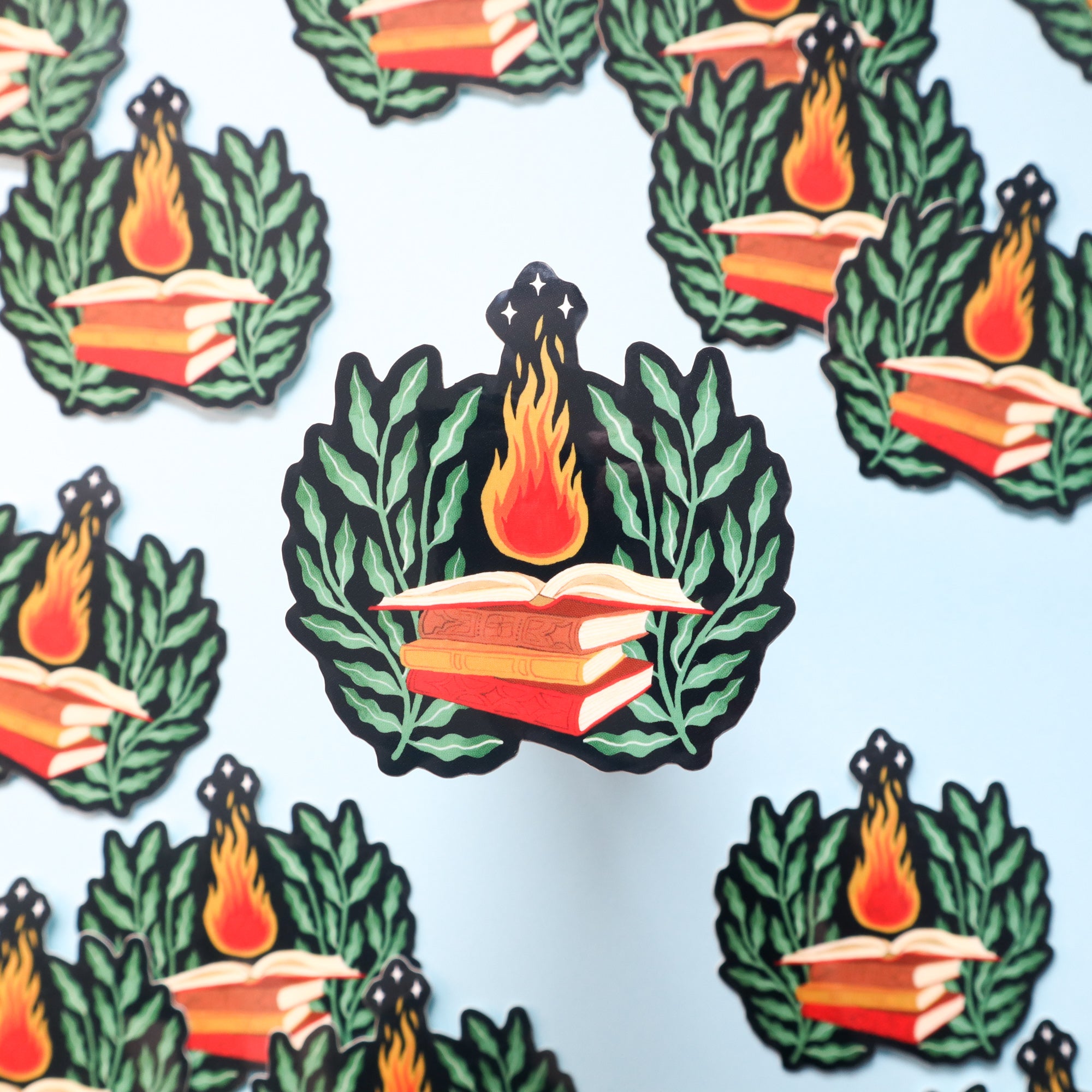 The Sorcerer Sticker by Papio Press features an open book with flames, green leaves, and stars on a light background. Each glossy vinyl sticker is UV-protected for vibrant, lasting color.