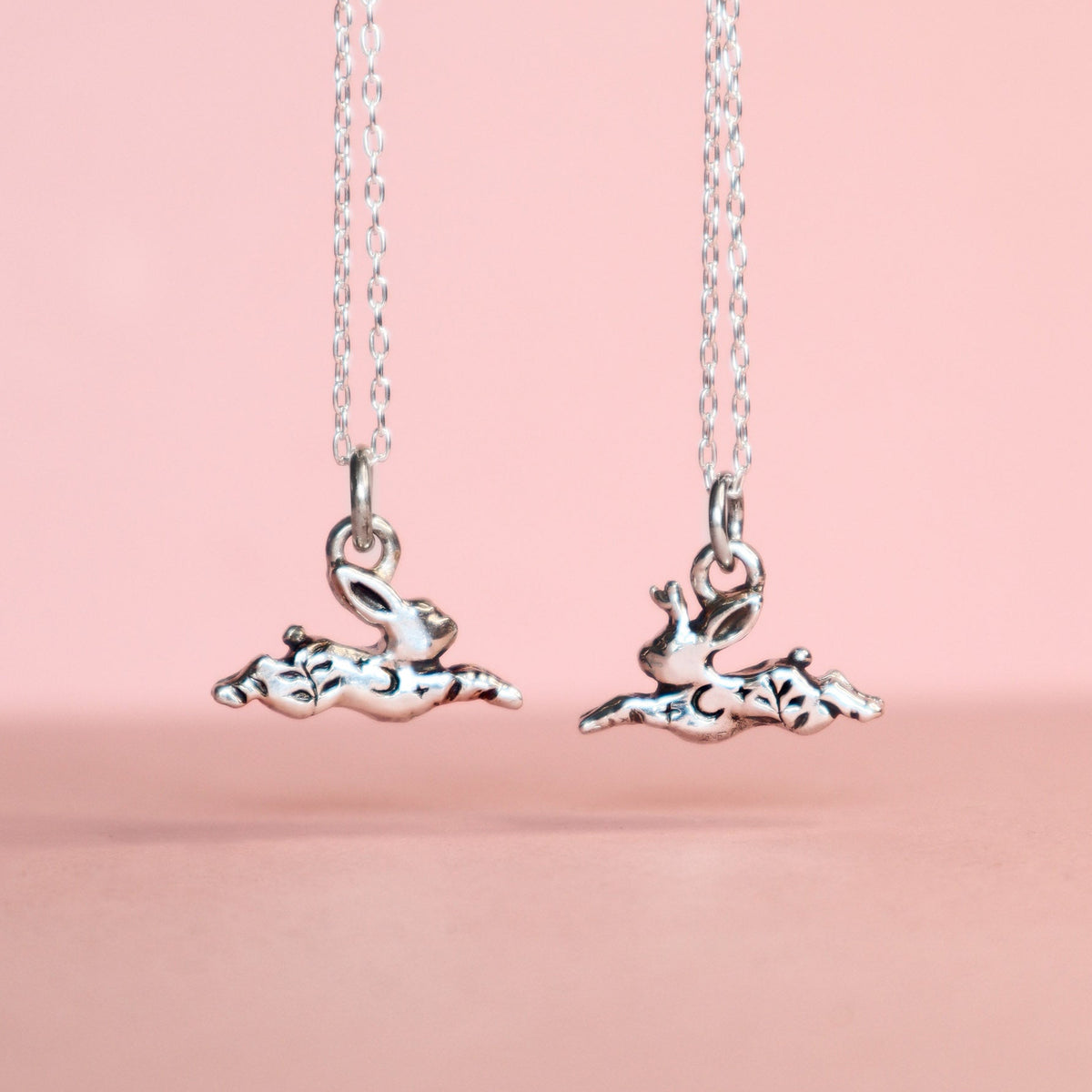 Two silver pendants, one rabbit and one jackalope hanging from cable link chains on a light pink background. The pendants have moon, star and leaf details.