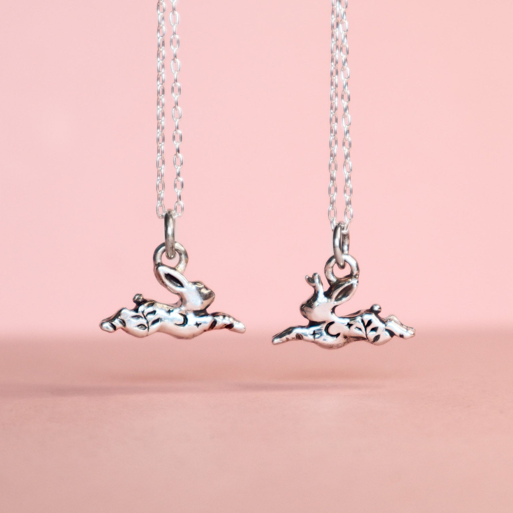 Two silver pendants, one rabbit and one jackalope hanging from cable link chains on a light pink background. The pendants have moon, star and leaf details.