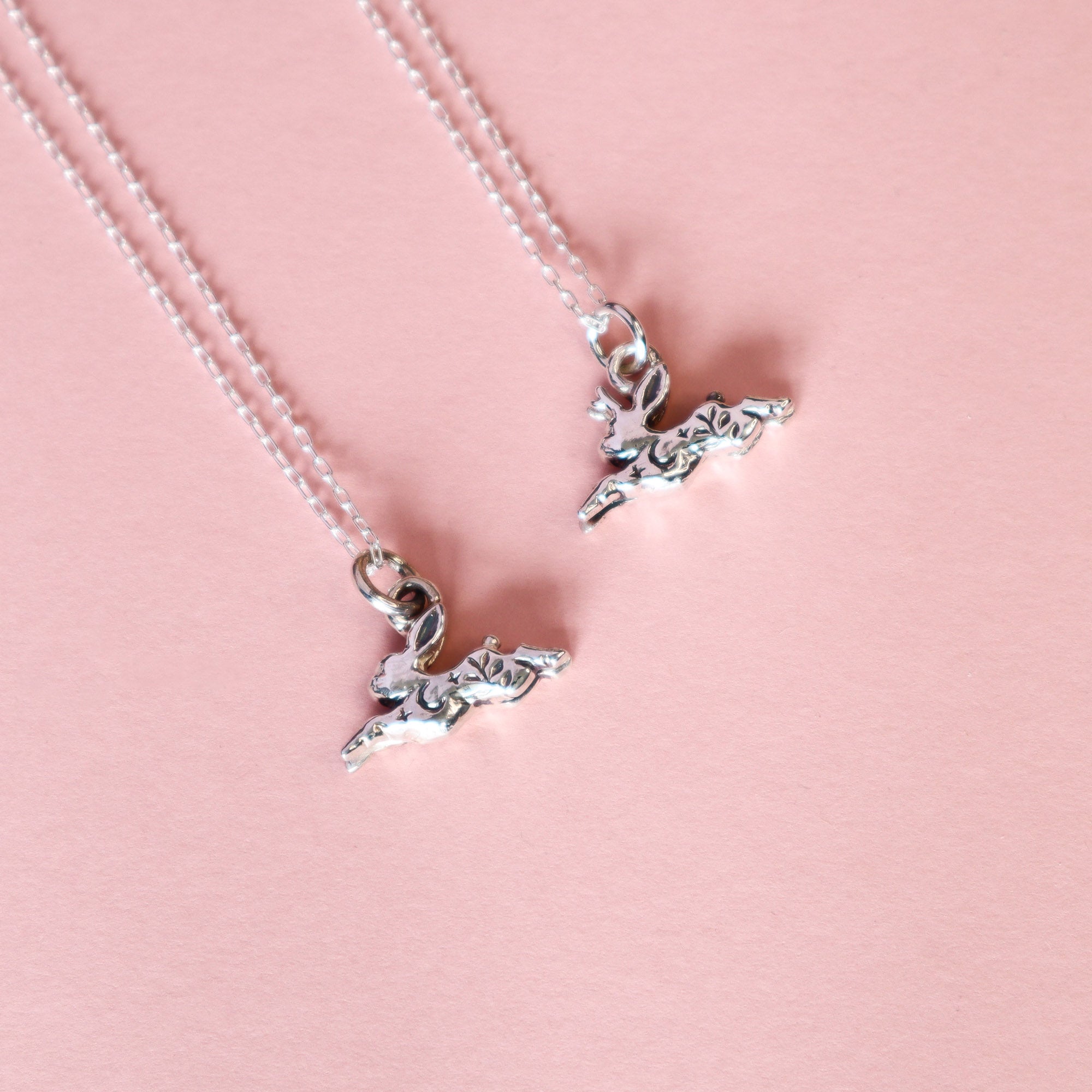 Two silver rabbit / jackalope pendants on a light pink background. 