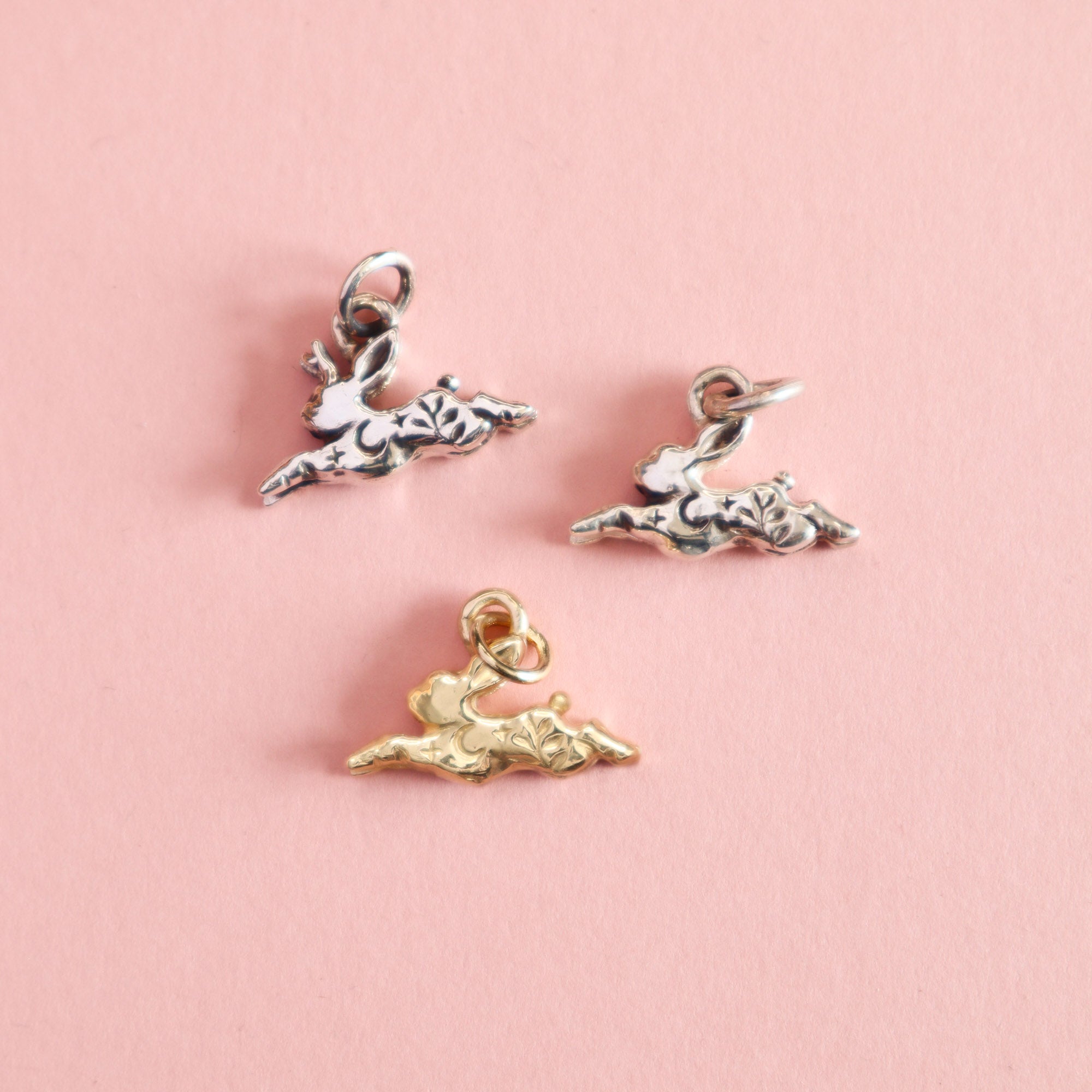 Three rabbit and jackalope charms in gold and silver on a pink background. The charms have moons and leaves engraved into them.