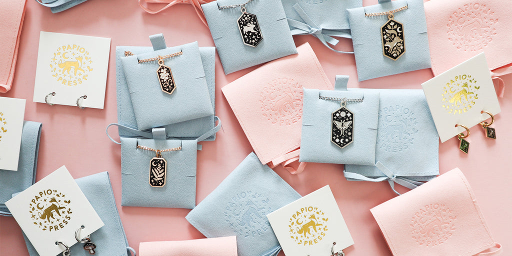 A photograph of a selection of Papio Press Jewellery, featuring necklaces and earrings on a pink background.