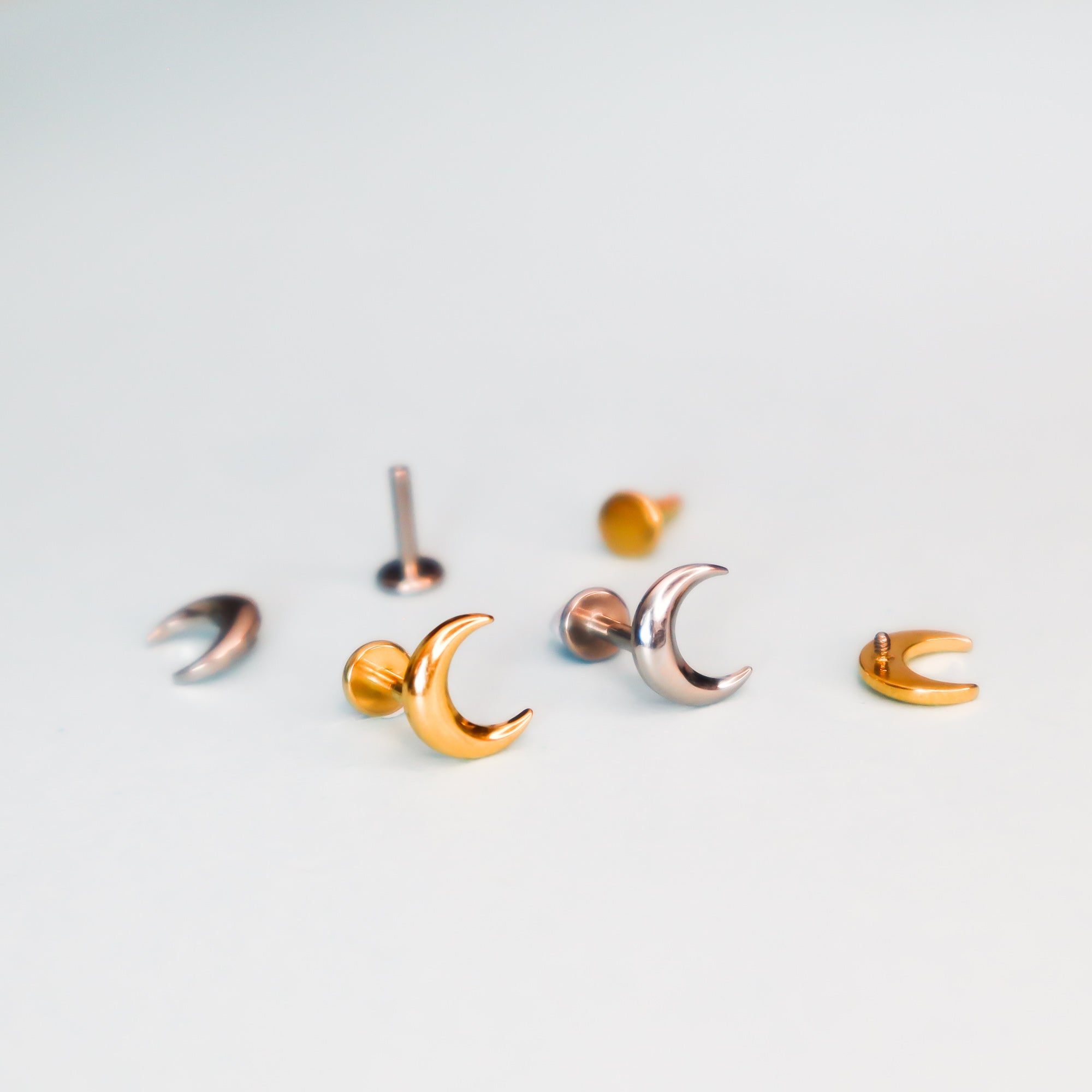 Papio Press Crescent Moon Labret Earrings in gold and silver tones, perfect for cartilage piercings, are arranged on a white surface with some lying flat and others upright.