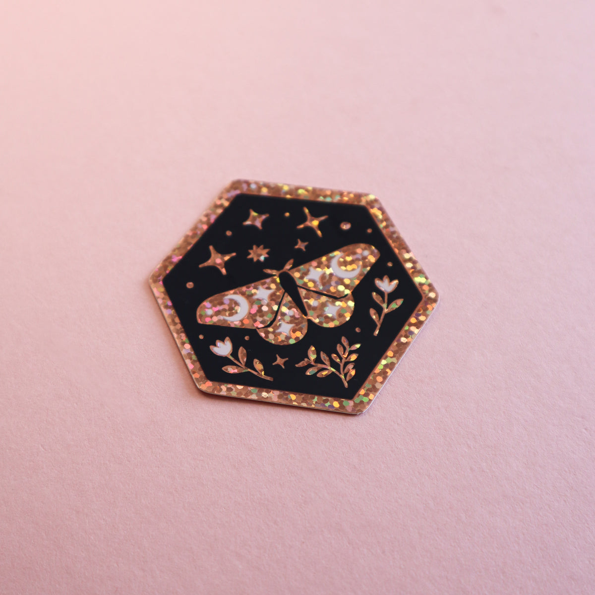 A hexagonal black sticker featuring a glittery gold butterfly, stars, and floral details, set against a soft pink background.