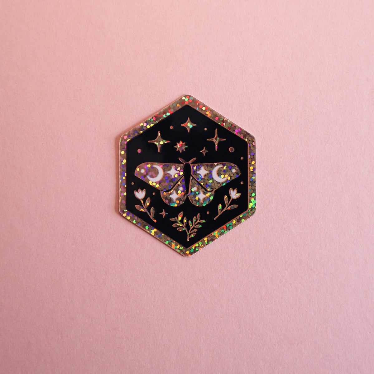 A hexagonal black sticker with a holographic glitter border and a central moth design, displayed on a solid pink background.