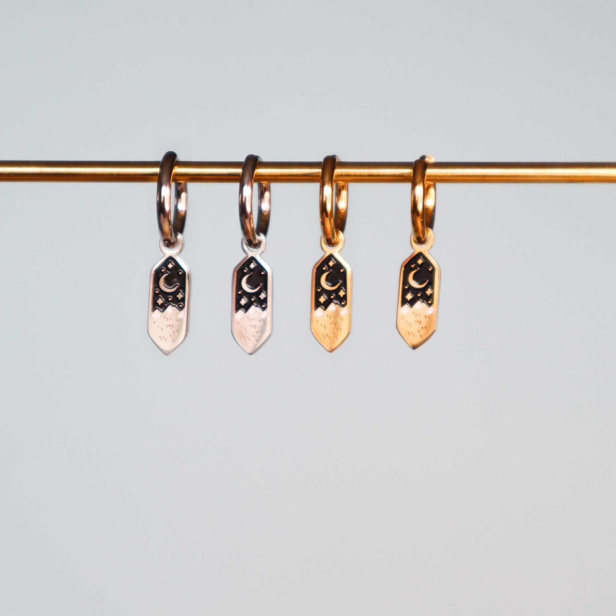 Four small hoop earrings, two silver and two gold, from Papio Press’s Mini Mountains collection, hang from a gold rod. Each features engraved crescent moons and stars on black enamel. A plain light gray backdrop completes the look.