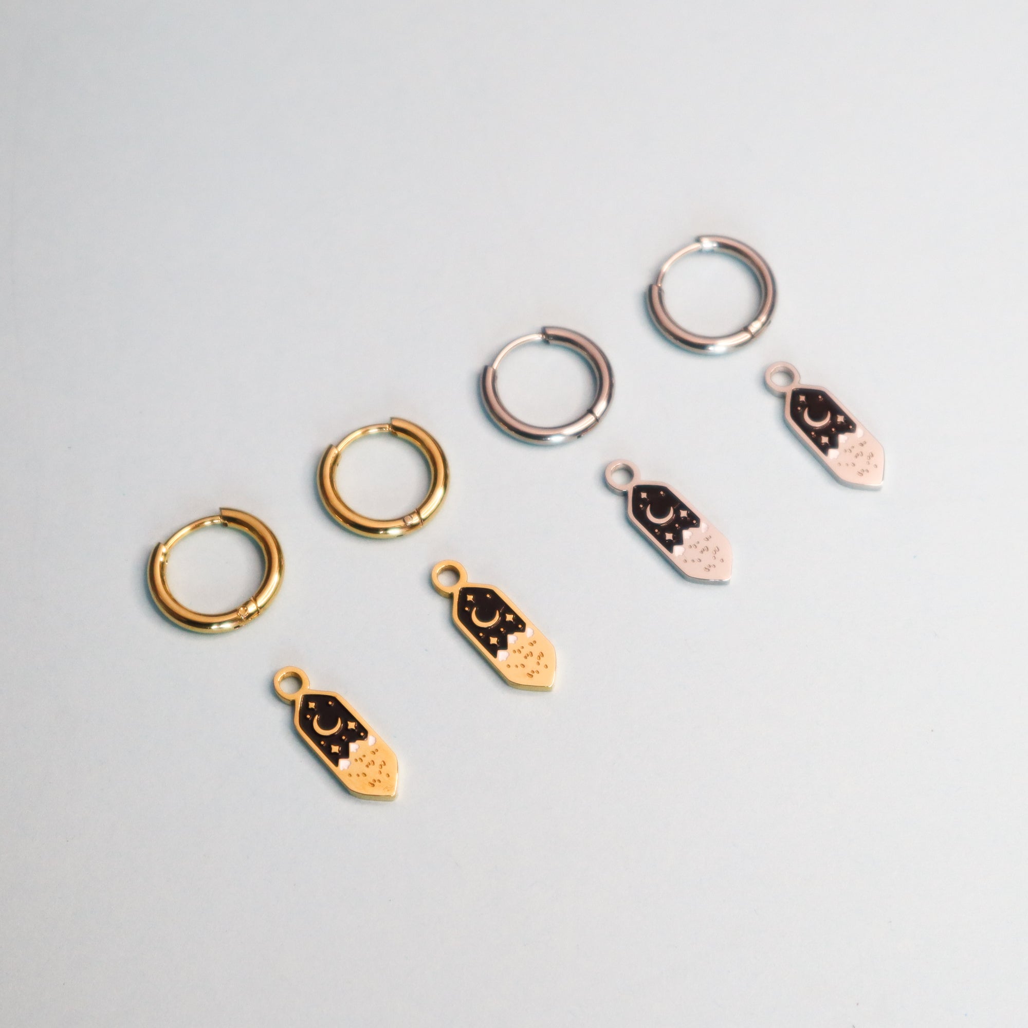 The Mini Mountains Hoop Earrings by Papio Press feature two gold plated and two silver small hoops, each paired with matching black enamel mountain scene charms, all displayed on a light gray background.