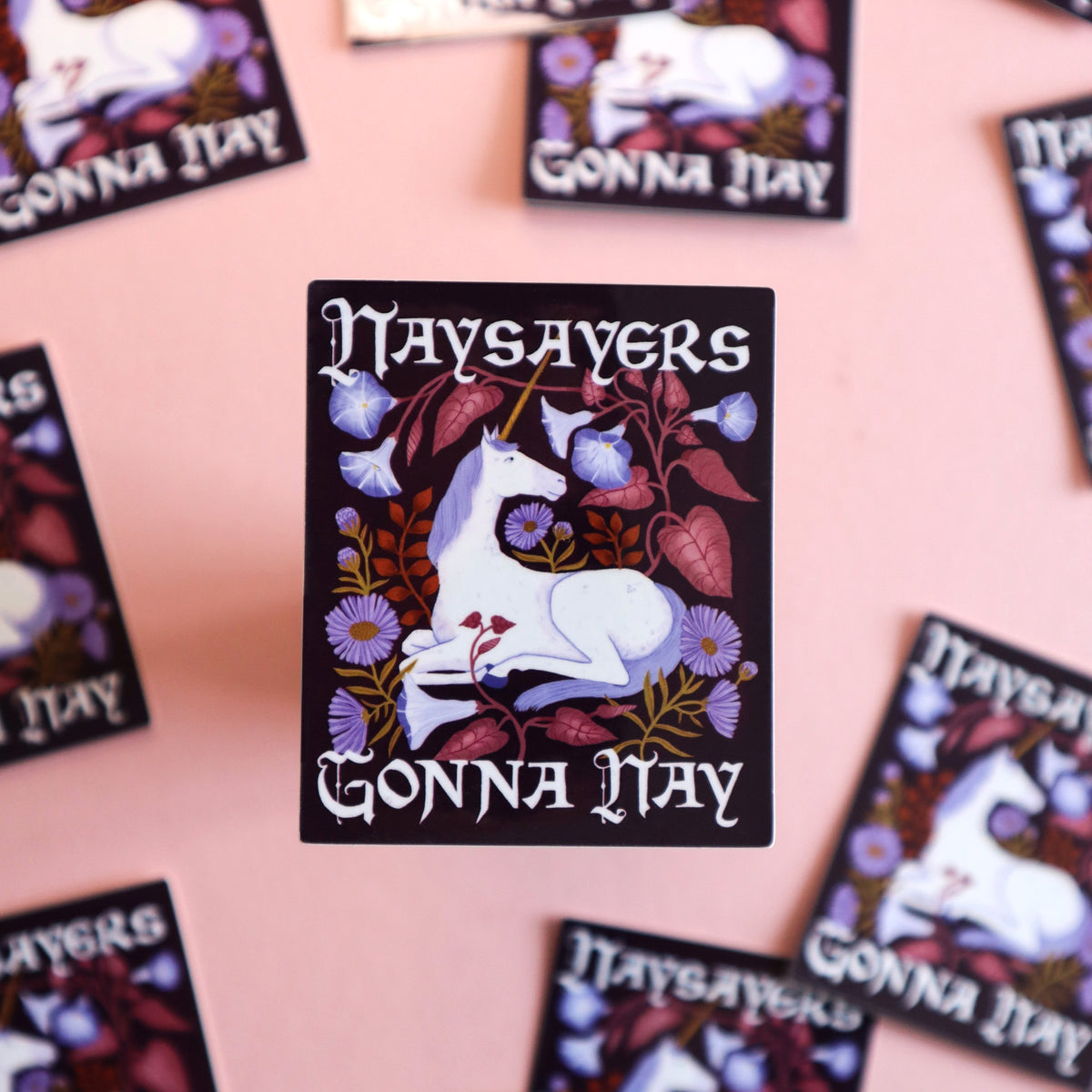 A square printed sticker centered on a pink surface, featuring a white unicorn surrounded by purple flowers and dark foliage, with the text 'Naysayers Gonna Nay'.