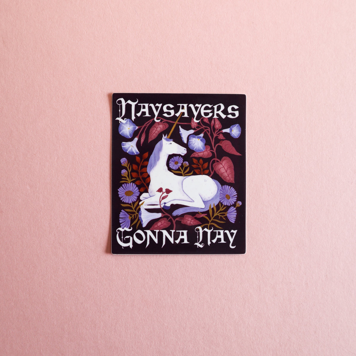 A square printed sticker centered on a pink surface, featuring a white unicorn surrounded by purple flowers and dark foliage, with the text 'Naysayers Gonna Nay'.