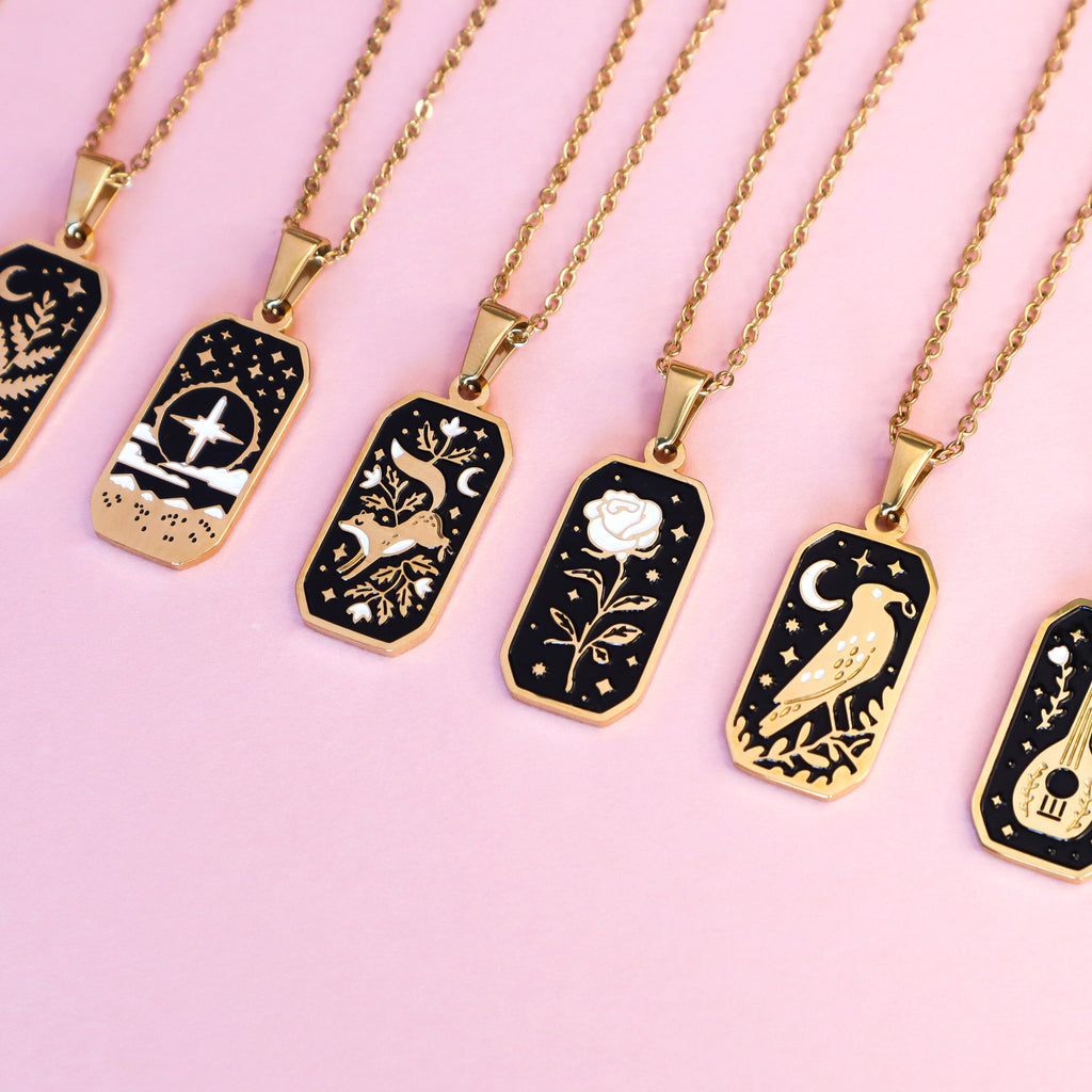A selection of 19K gold-plated stainless steel Papio Press necklaces. Featuring The Cleric, Floral Fox, Rose, The Crow and The Bard on a light pink background. 