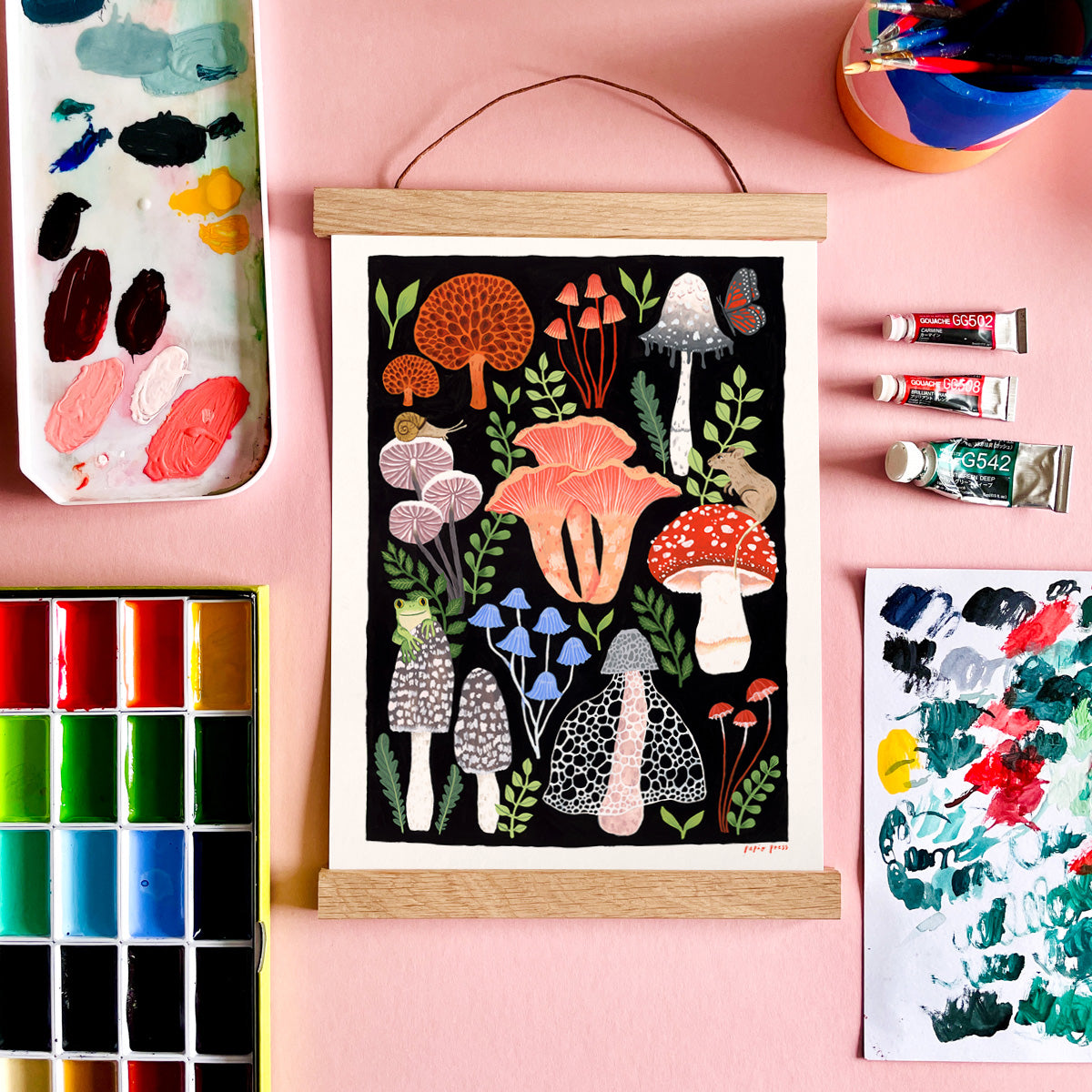 A Mushroom Papio Press print in a hanging frame surrounded by paints and palettes on a pink background. 