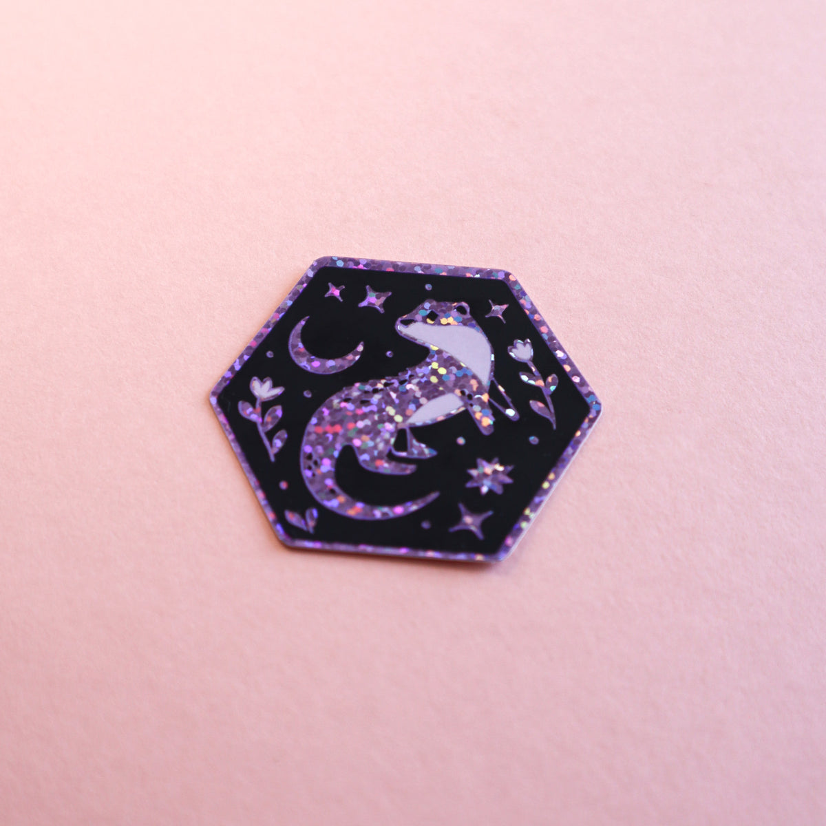 A hexagonal sticker or patch featuring a glittery purple and pink dragon on a black background, surrounded by stars and a crescent moon, set against a solid pink background.