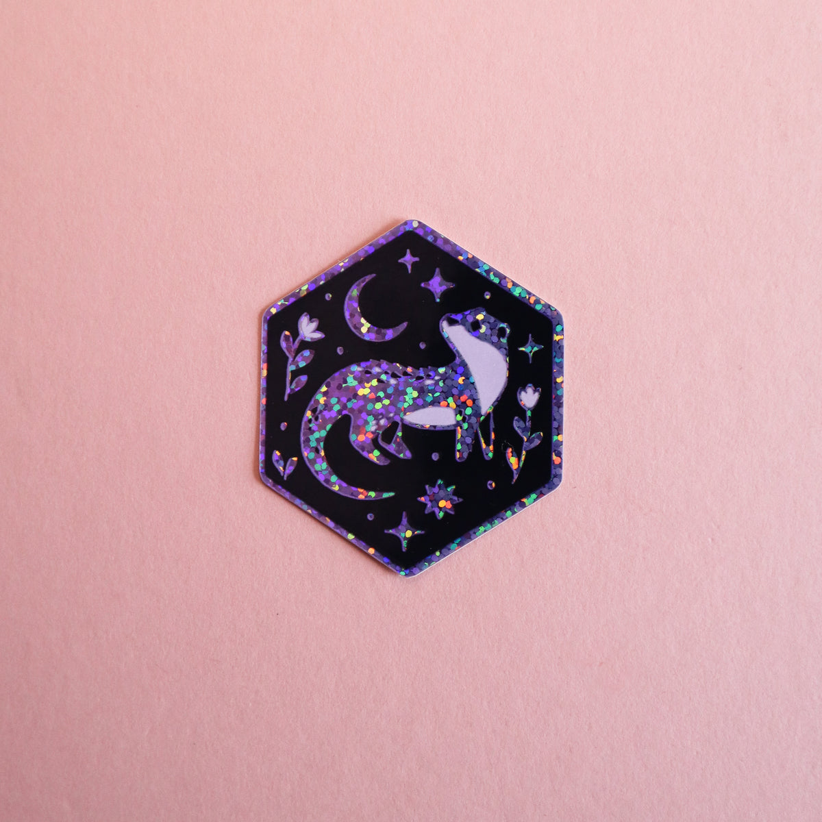 A hexagonal sticker featuring a glittery purple otter howling at a crescent moon and stars on a black background, placed on a solid pink surface.
