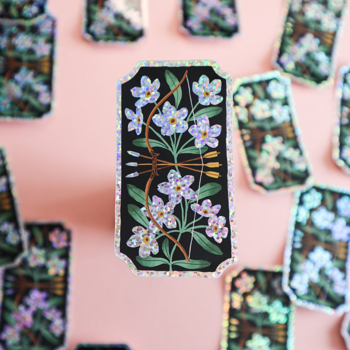 A rectangular sticker with a black background, decorated with blue forget-me-not flowers, green leaves, and a brown bow and arrow, finished with a holographic glitter border, set against a solid pink background.