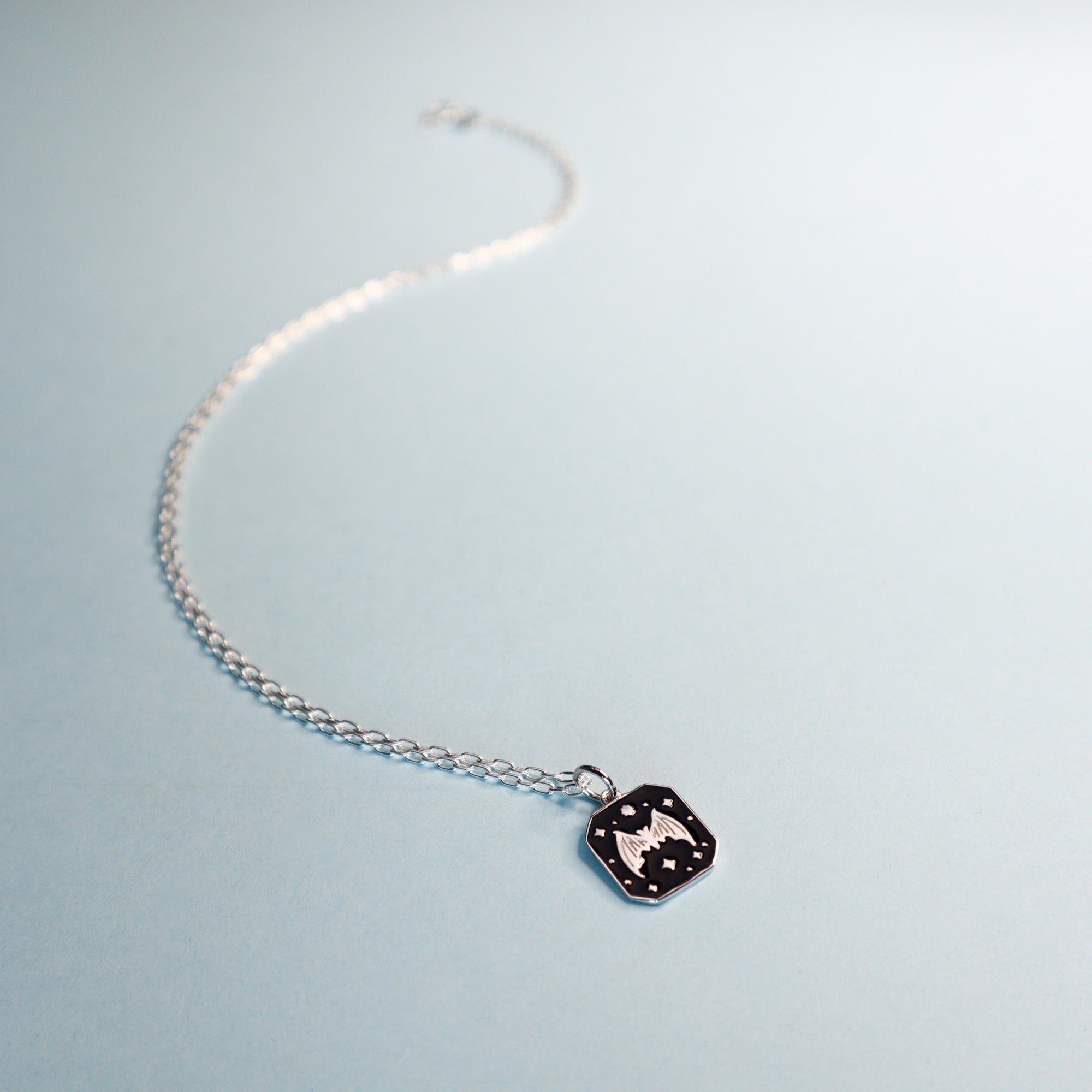 Square silver pendant featuring a Bat and stars with black enamel details. Taken on a light blue background.
