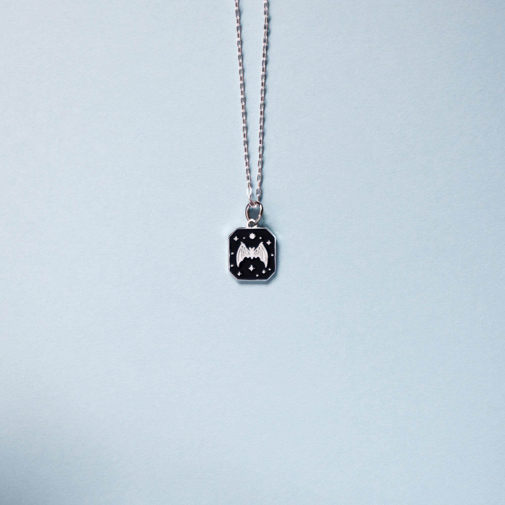 Square silver pendant featuring a Bat and stars with black enamel details. Taken on a light blue background.