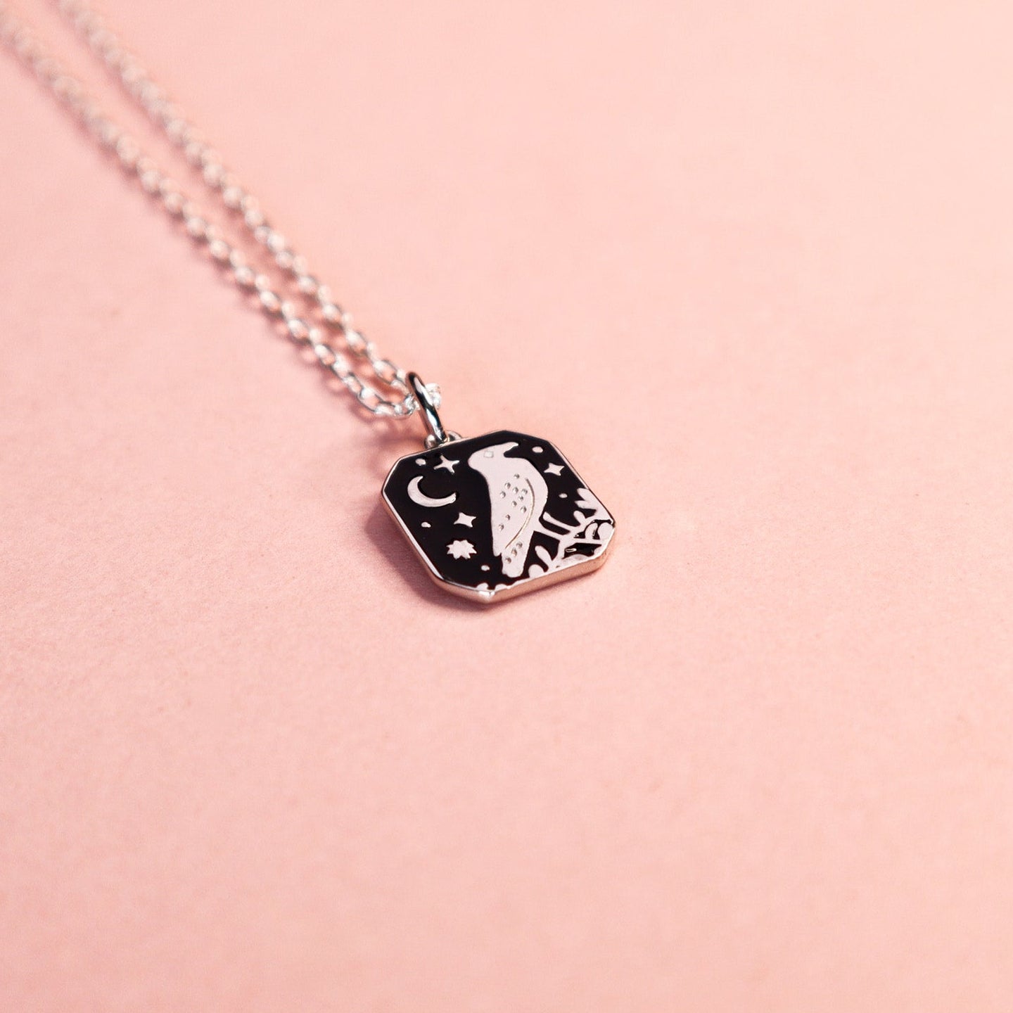 Square silver pendant featuring a crow, moon and stars with black enamel details. Taken on a light pink background.
