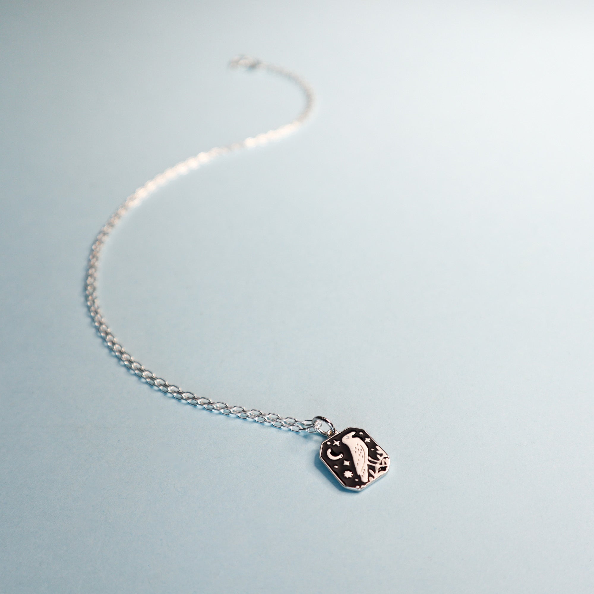 Square silver pendant featuring a crow, moon and stars with black enamel details. Taken on a light blue background.