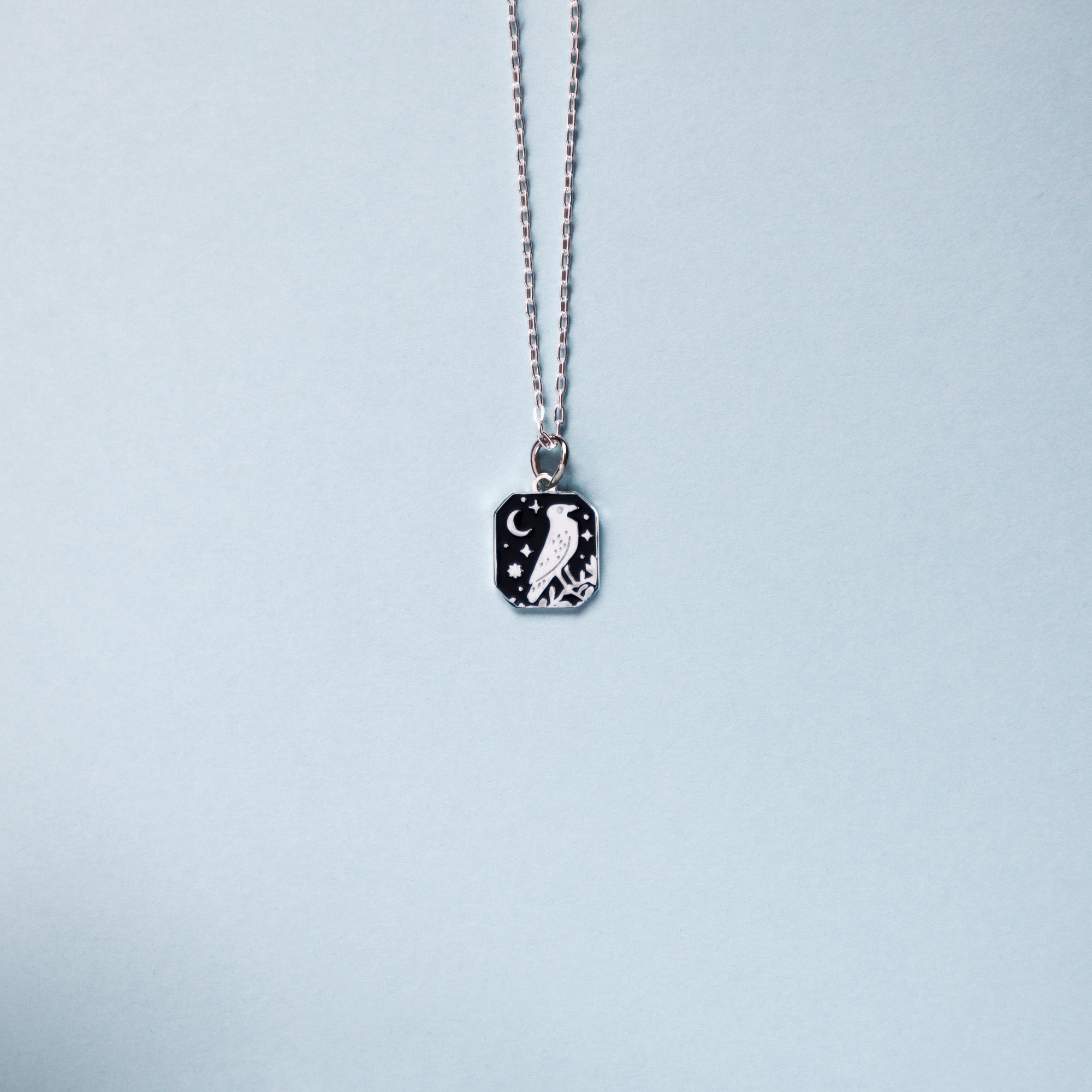 Square silver pendant featuring a crow, moon and stars with black enamel details. Taken on a light blue background.