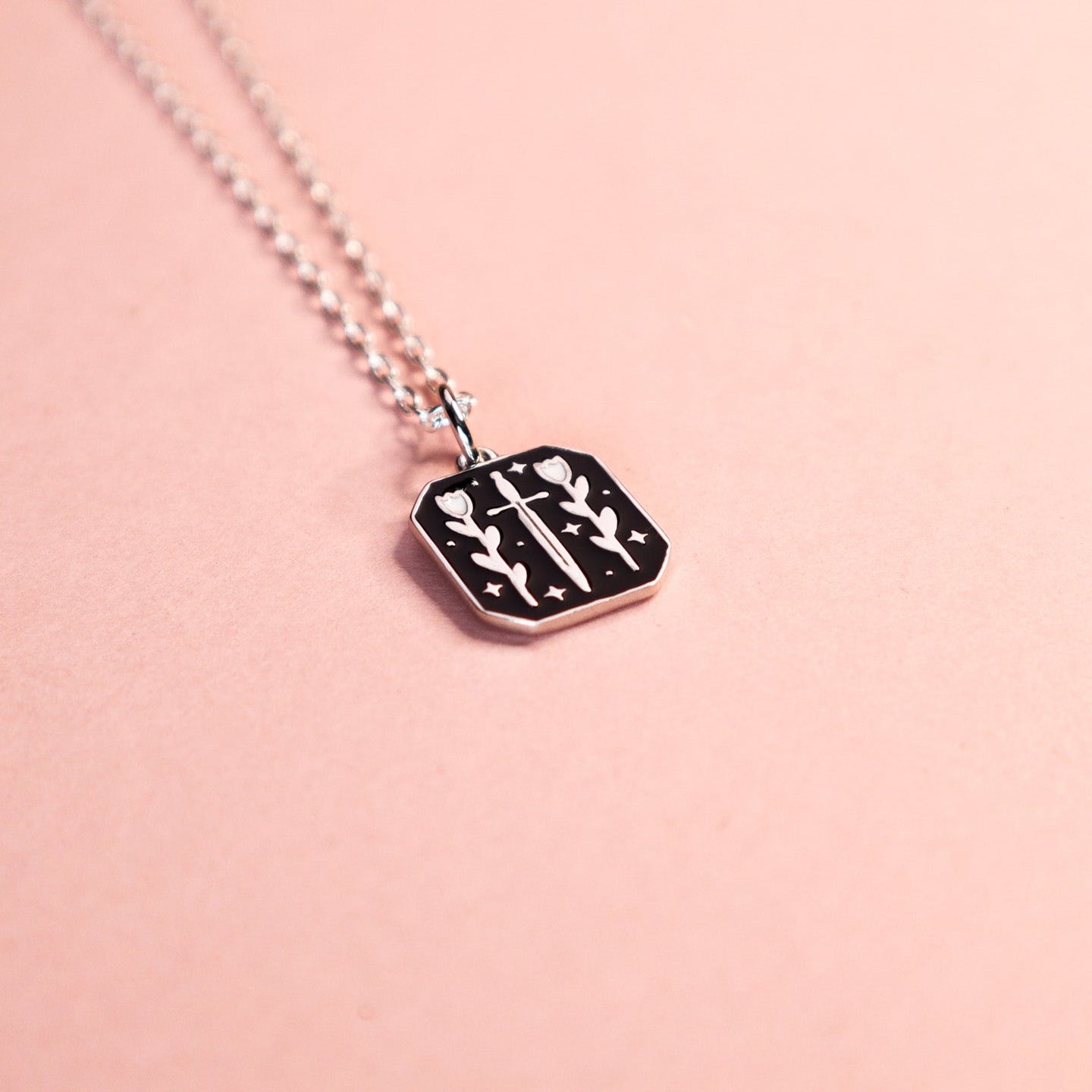 Square silver pendant featuring a dagger, florals and stars with black enamel details. Taken on a light pink background.