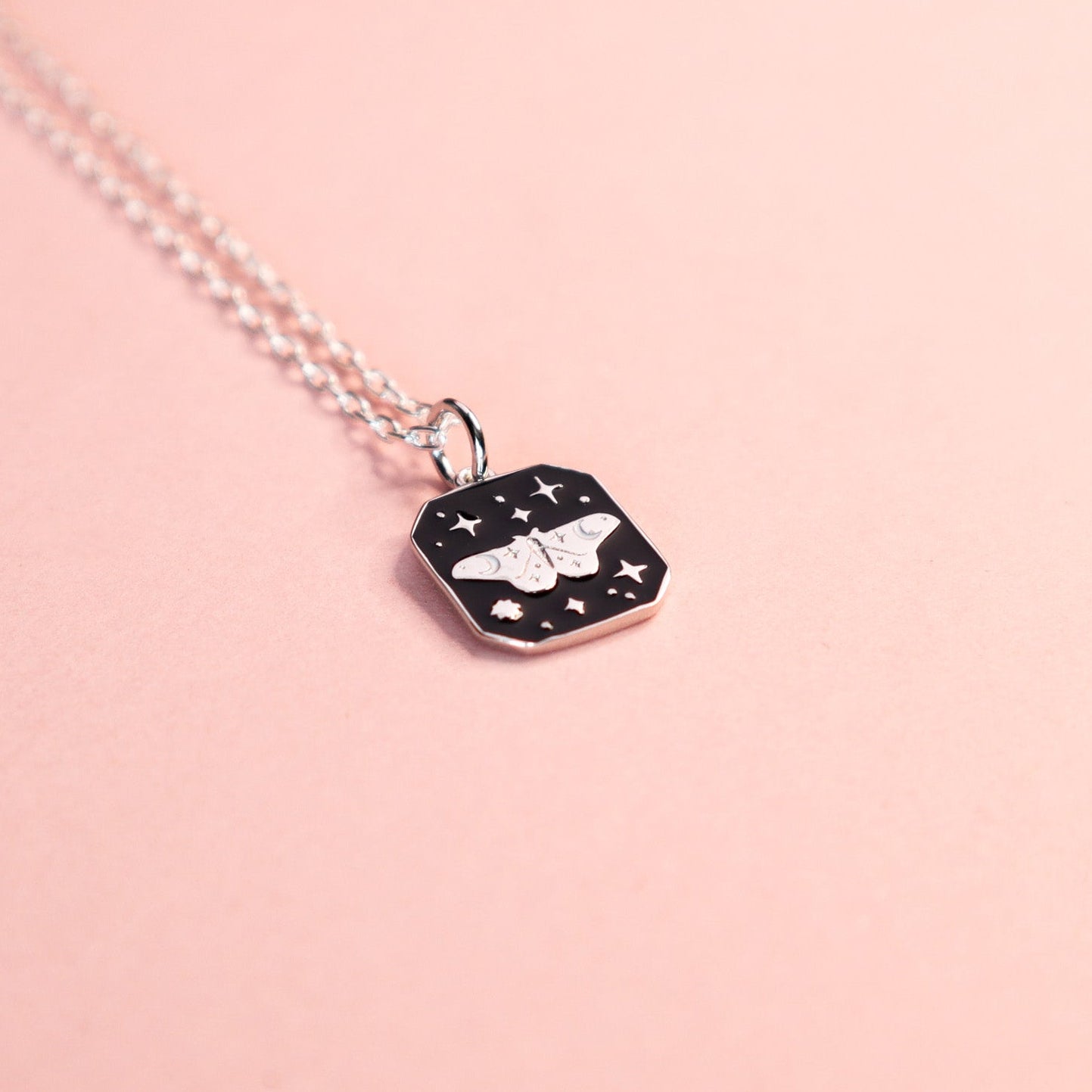 Square silver pendant featuring an moth and stars with black enamel details. Taken on a light pink background.
