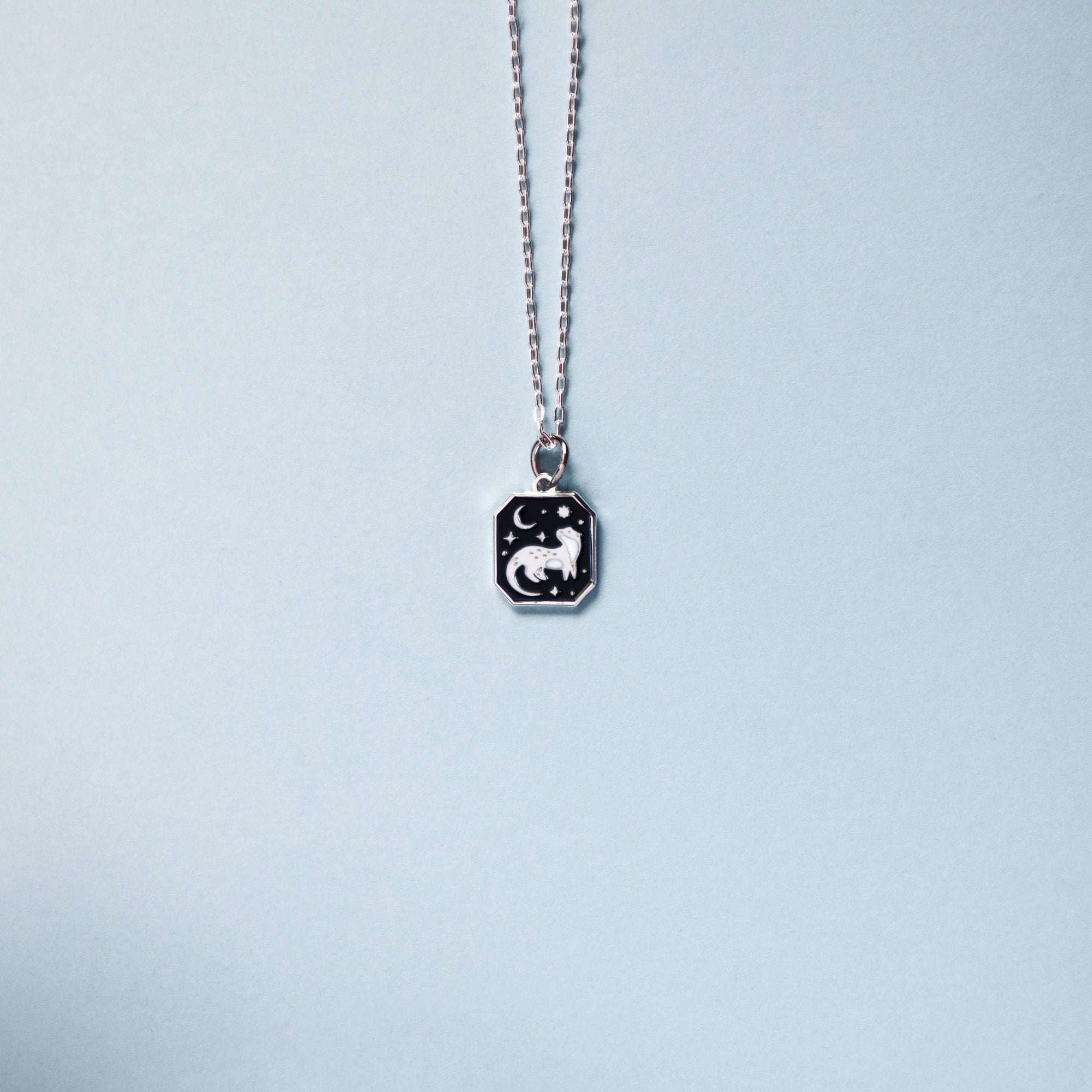 Square silver pendant featuring an otter, moon and stars with black enamel details. Taken on a light blue background.