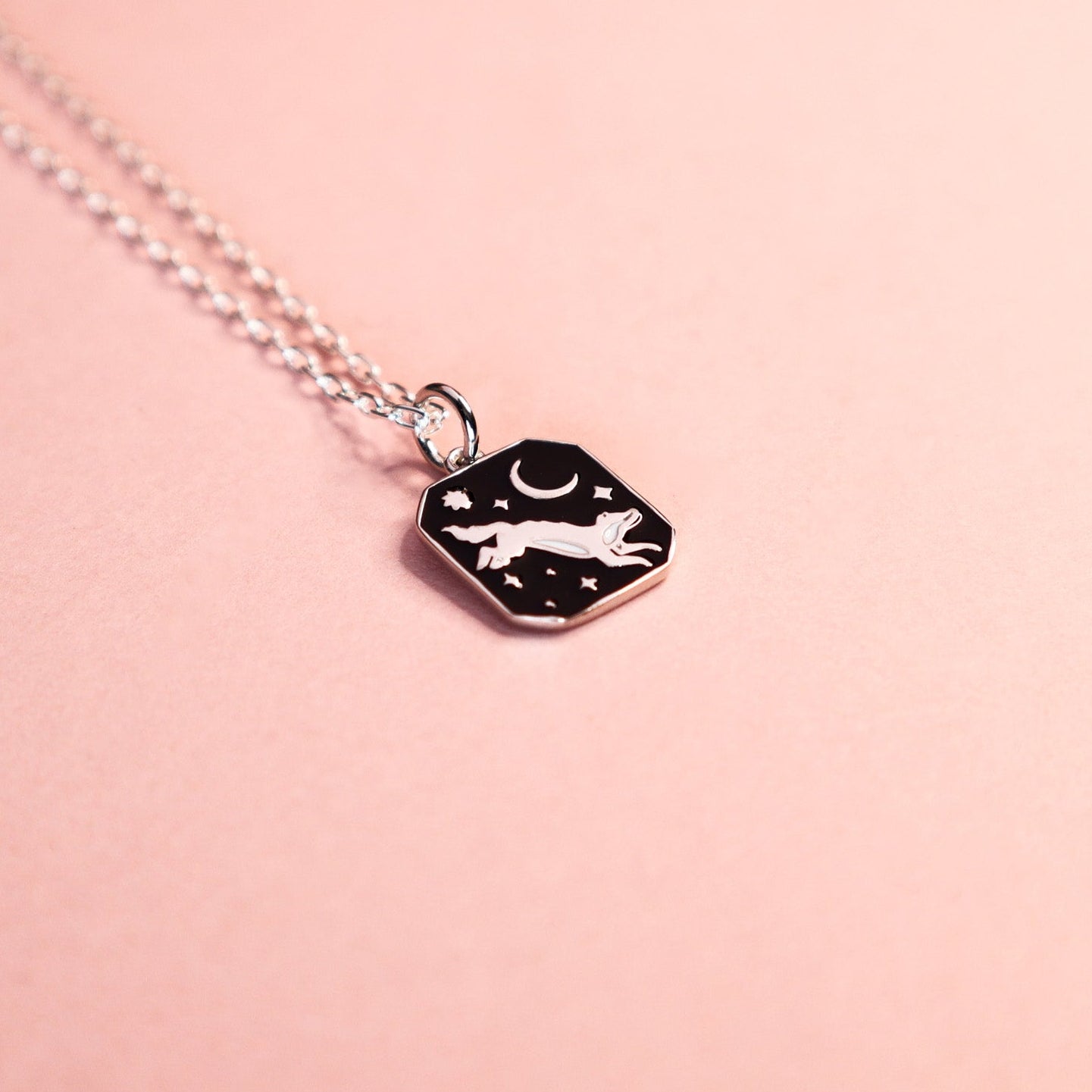 Silver necklace with a black pendant featuring a wolf, moon and star design on a pink background