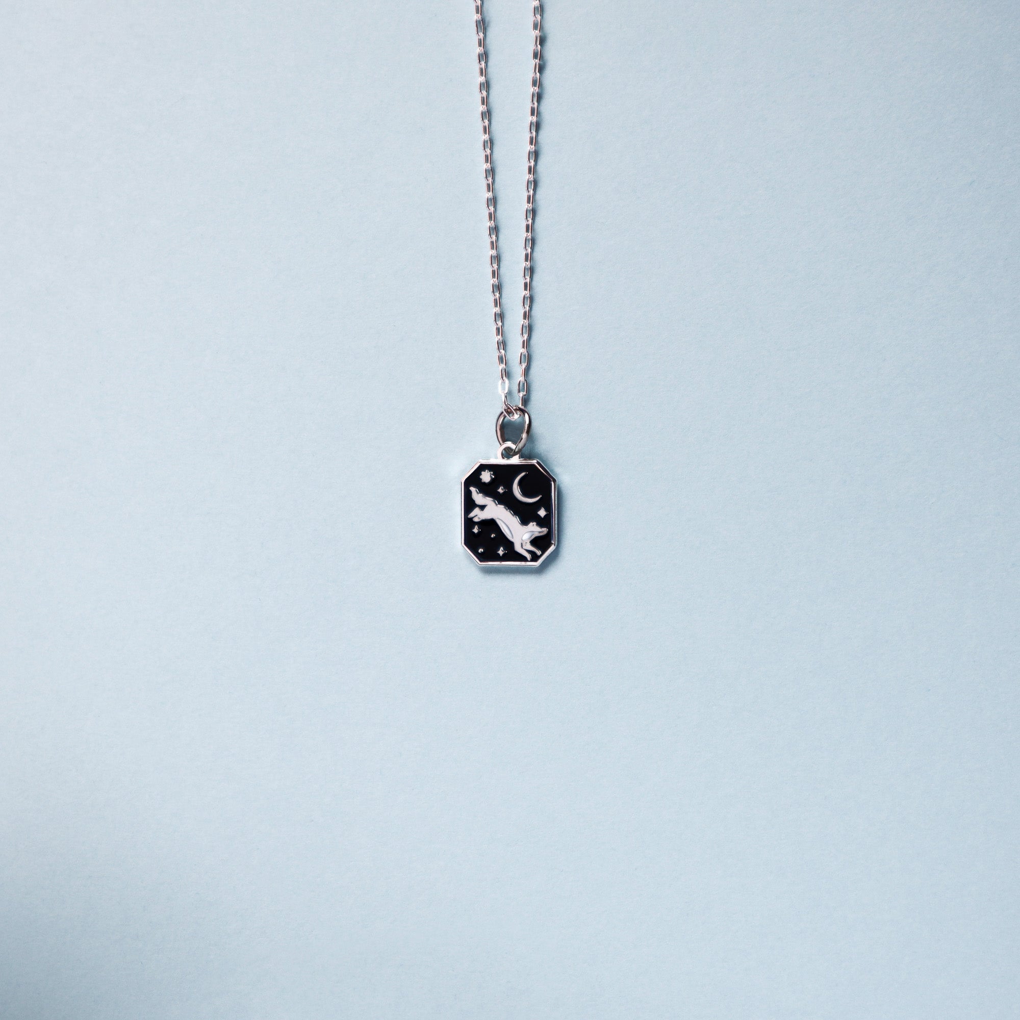 Square silver pendant featuring an wolf, moon and stars with black enamel details. Taken on a light blue background.