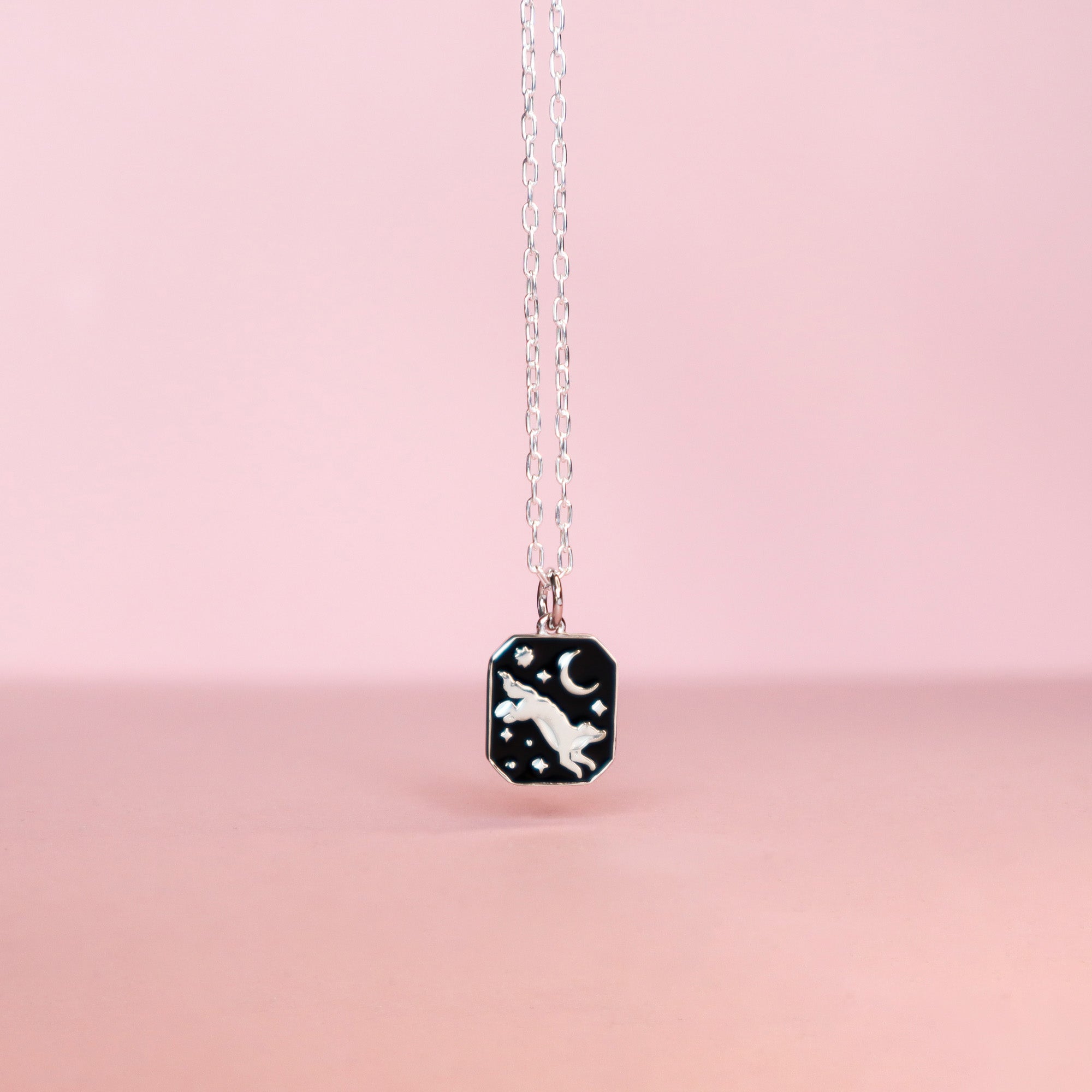 A hanging square silver pendant featuring an wolf, moon and stars with black enamel details. Taken on a light pink background.