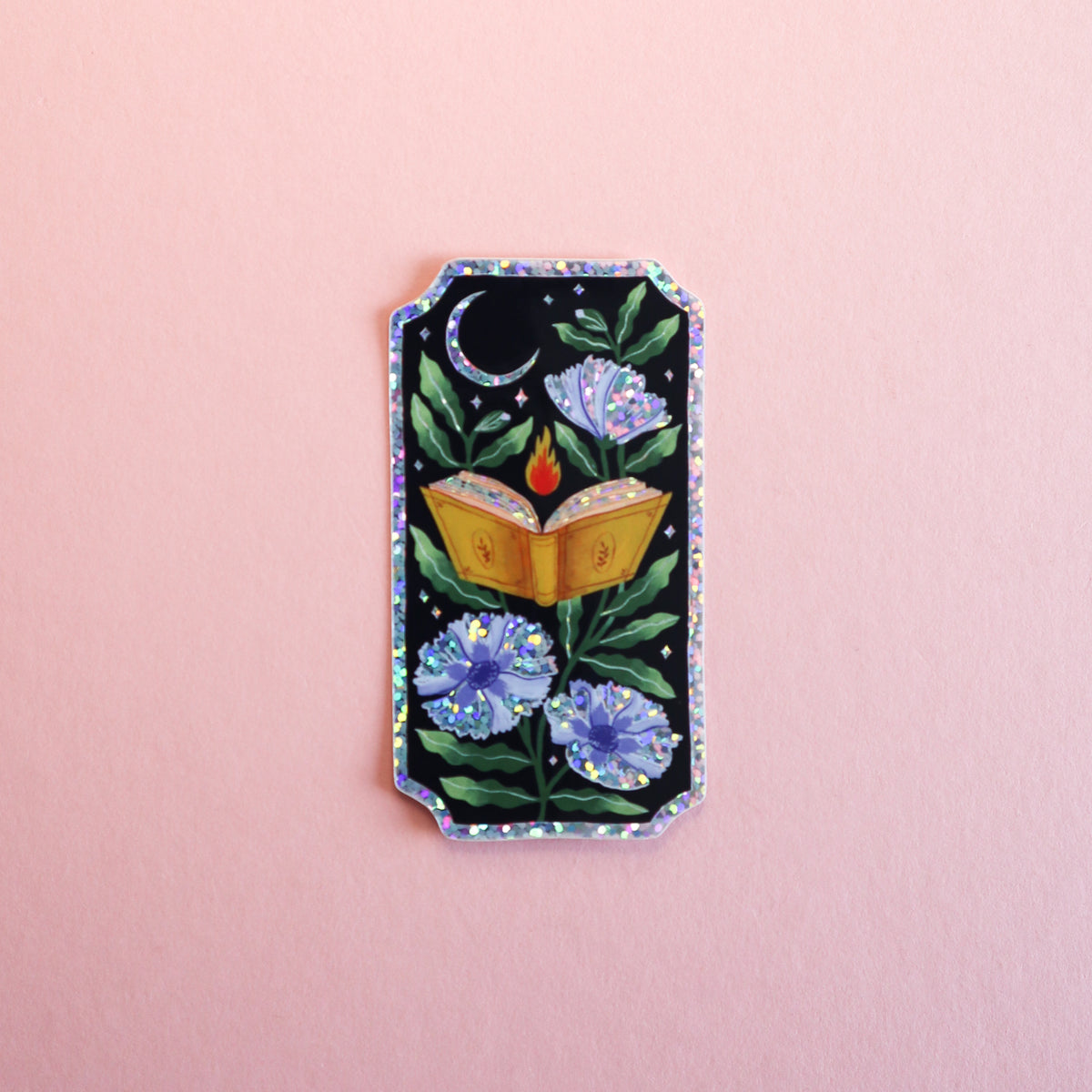 A rectangular sticker with a black background, decorated with blue flowers, green leaves, and an open book, finished with a holographic glitter border, set against a solid pink background.