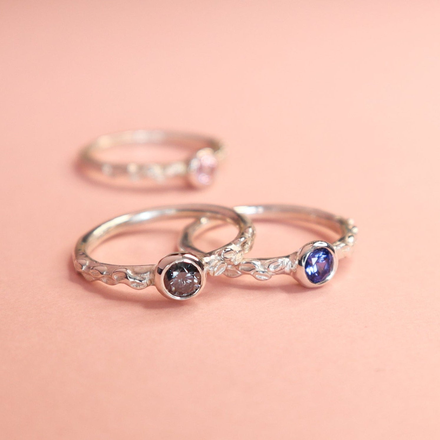 Three Silver rings on a light pink background. Each has a different stone, one blue sapphire, one pink sapphire and one grey moissanite. 