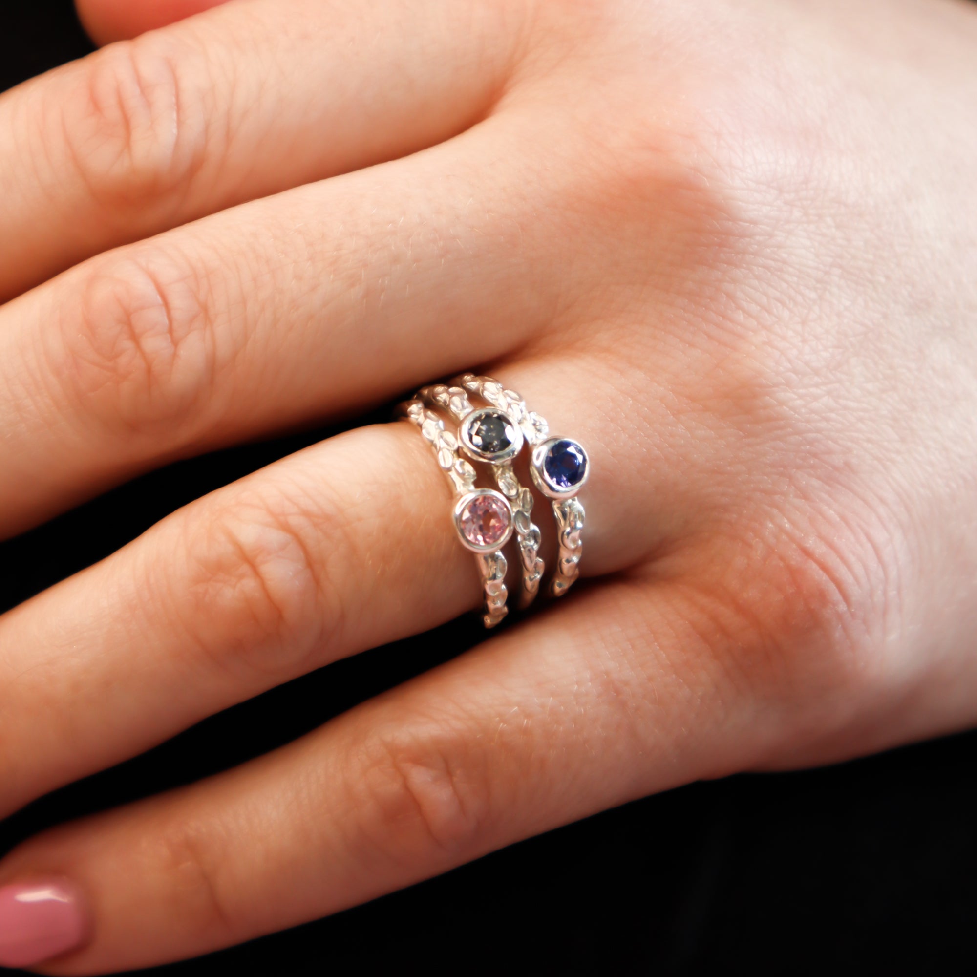 Three silver rings stacked on hand against a black background. One ring features a blue sapphire, one a pink sapphire and the other a grey moissanite.