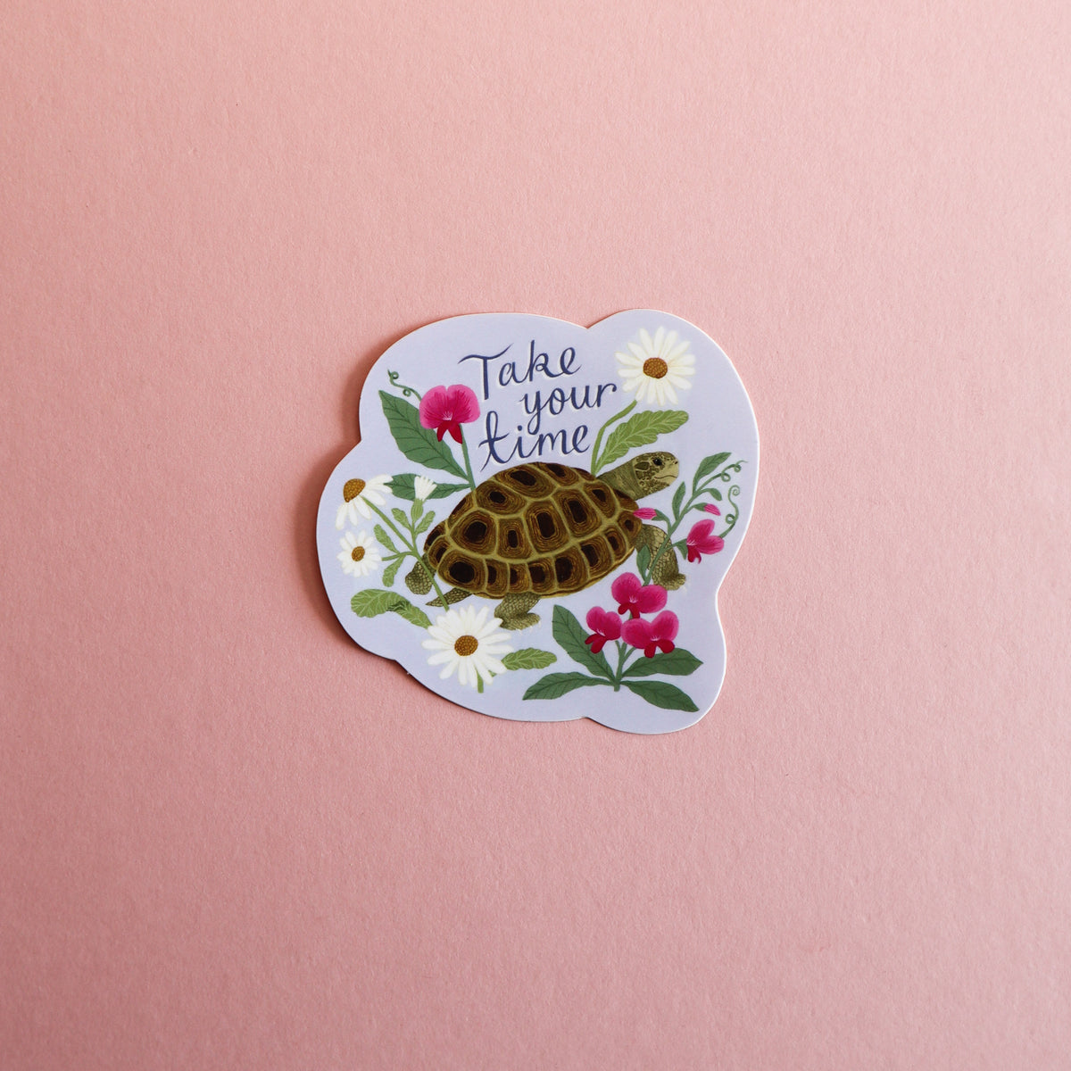 A die-cut sticker featuring a tortoise surrounded by white daisies and pink flowers with the text 'Take your time' in black script, displayed on a solid pink background.