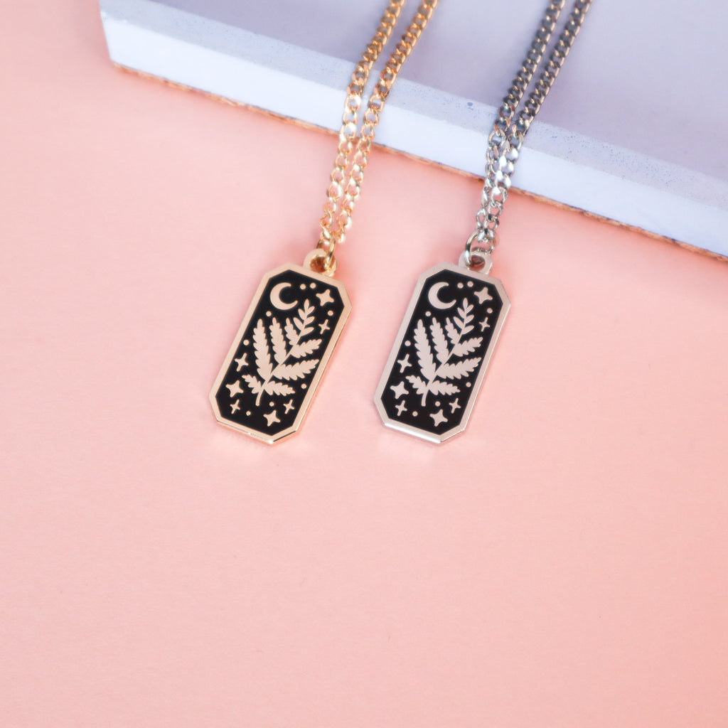 A photograph of two necklaces, one silver, one gold. The necklaces are rectangular and feature a leaf, moon and stars.