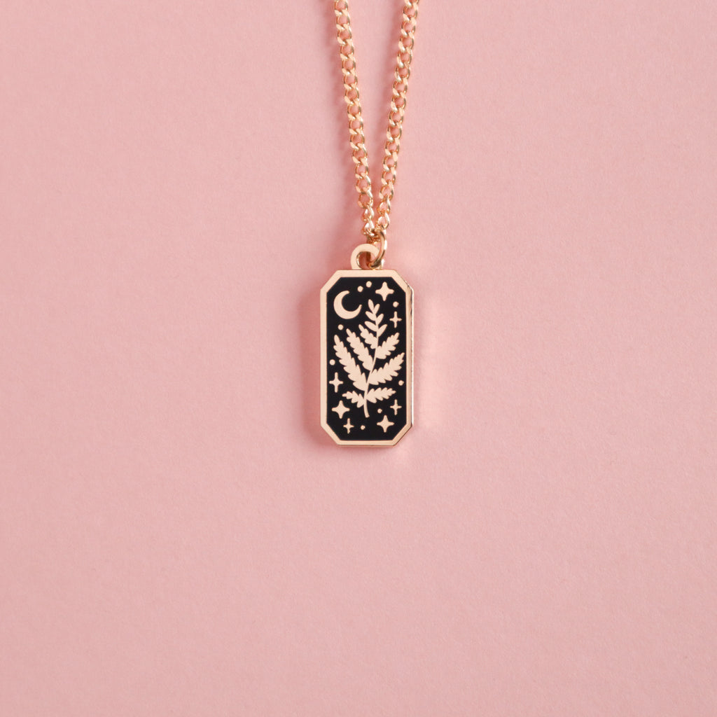A photograph of a gold plated necklace. The necklaces are rectangular and feature a leaf, moon and stars and is taken on a pink background.
